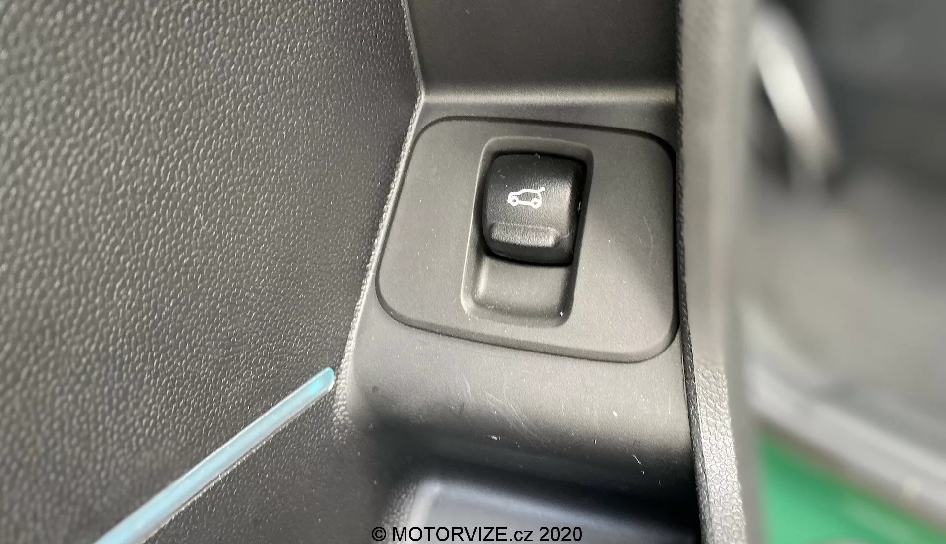 Close-up do interruptor de controle para operar os espelhos laterais no interior do Mini Countryman 2020 John Cooper Works. O interruptor retangular com cantos arredondados, localizado em um painel plano, é destacado com um símbolo de espelho. Um lado do interruptor está na sombra, indicando uma fonte de luz proveniente do lado oposto. O fundo apresenta plástico preto texturizado e uma fina linha azul de iluminação ambiente, contribuindo para a estética interior do veículo.