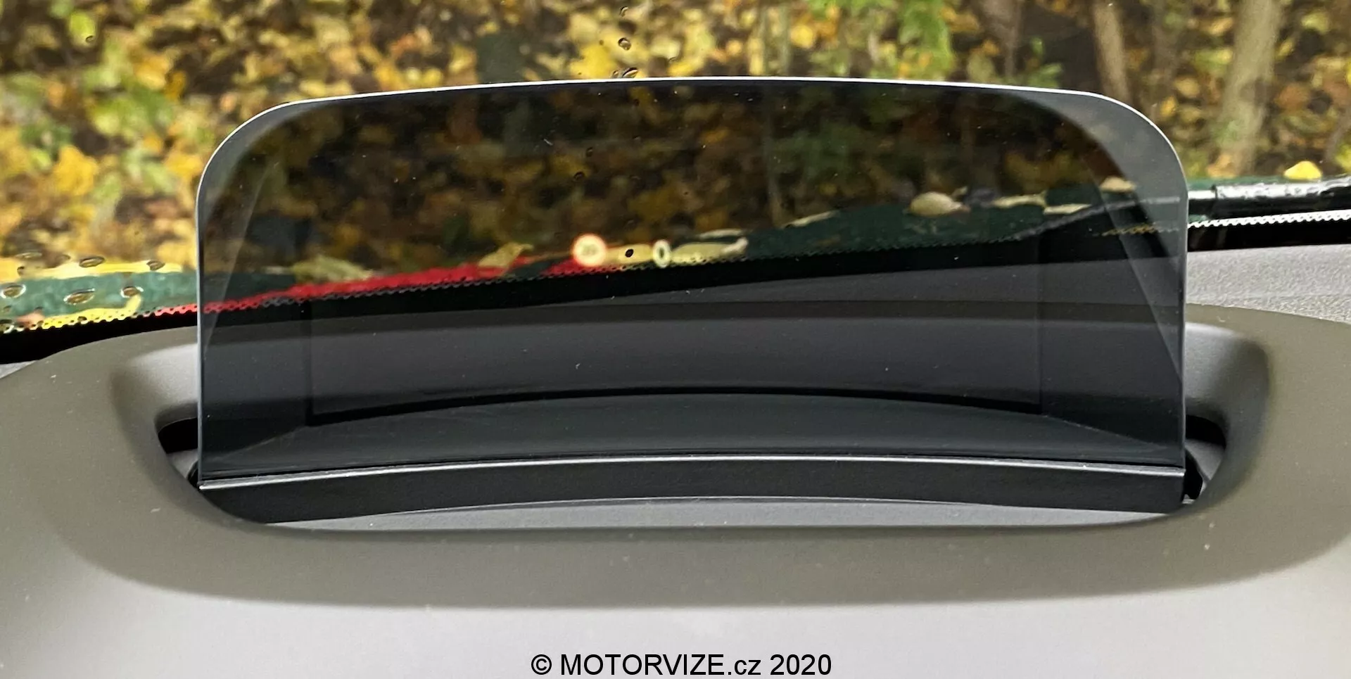 Imagem mostrando o head-up display (HUD) do Mini Countryman 2020, projetado em uma tela transparente acima do painel a partir da perspectiva do motorista. O painel HUD claro e semelhante ao vidro é perfeitamente integrado ao design interior, arredondado e ligeiramente inclinado para visibilidade ideal. O fundo fora do para-brisa está desfocado, enfatizando o foco do HUD em apresentar informações de condução cruciais.