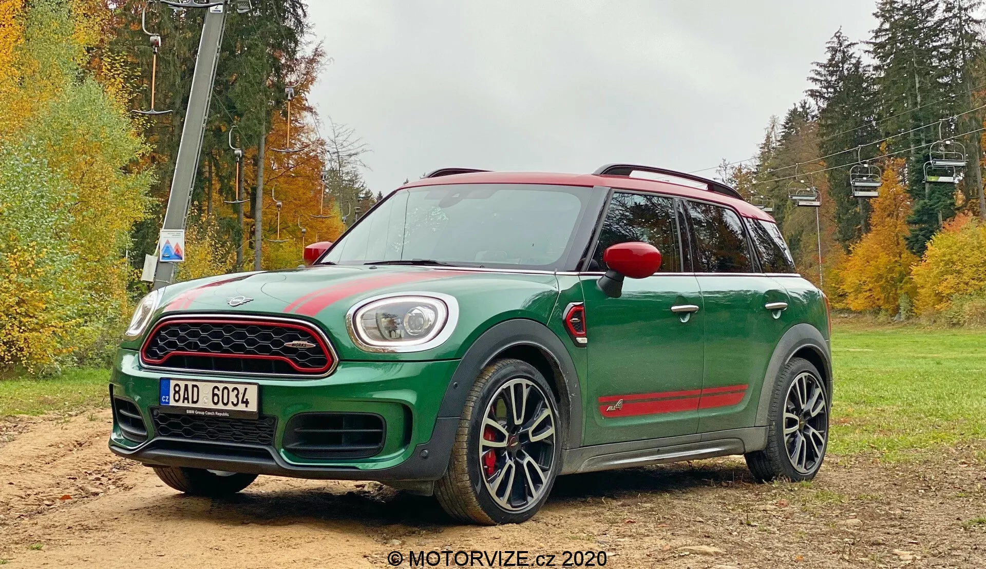 Imagem de um Mini Countryman John Cooper Works 2020 verde em uma área de cascalho off-road com uma paisagem florestal ao fundo. O crossover compacto exibe uma visão frontal de três quartos com teto e espelhos laterais vermelhos contrastantes, grande grade frontal com acabamento cromado, luzes diurnas em LED que enquadram os faróis principais e luzes de nevoeiro integradas no para-choque inferior. As características esportivas notáveis incluem uma tomada de ar no capô, revestimentos pretos dos arcos das rodas, saias laterais, spoiler traseiro, grandes rodas de liga leve de múltiplos raios com pneus de perfil baixo e pinças de freio vermelhas. Também são visíveis um teto solar panorâmico e barras de teto aerodinâmicas.