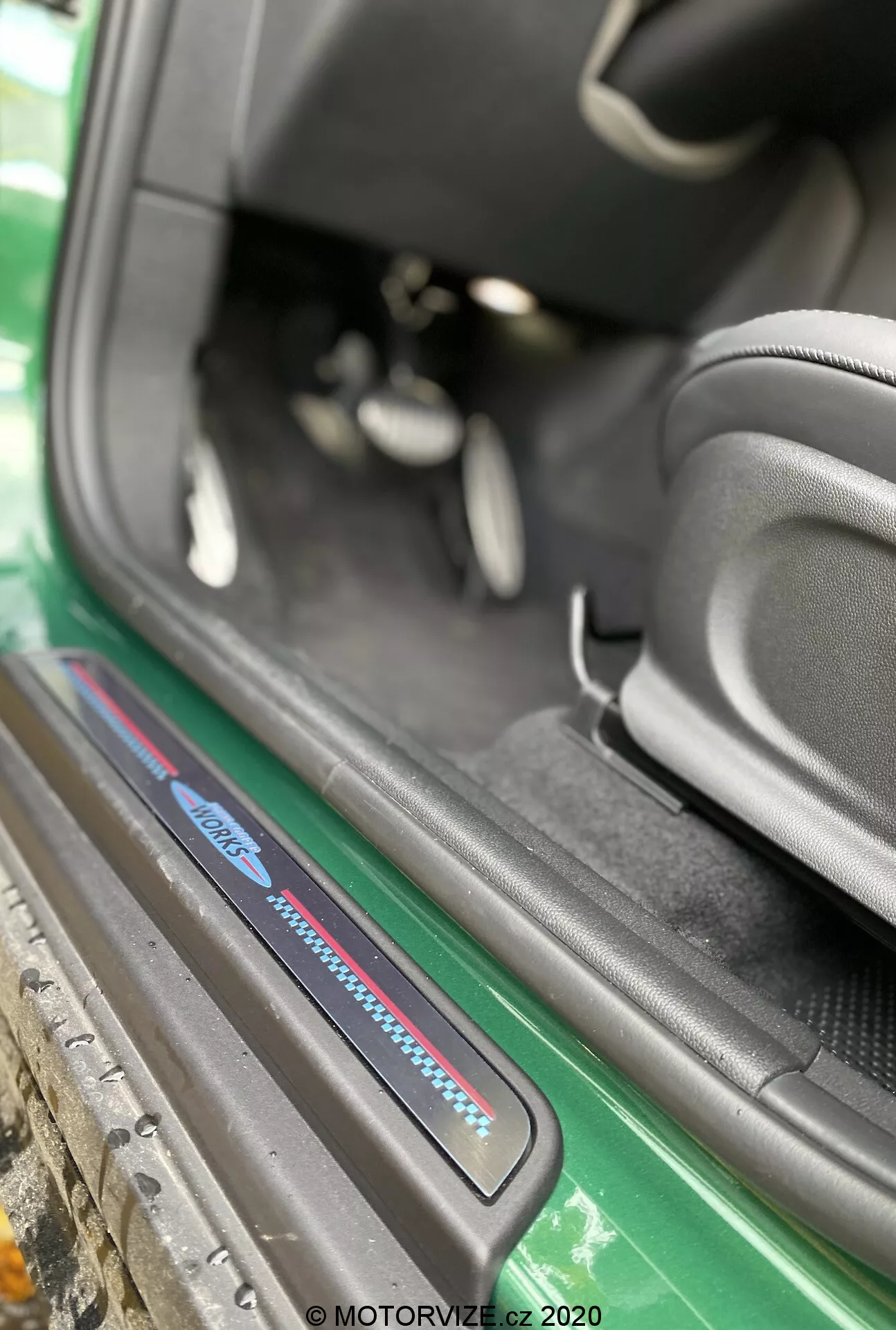 Vista aproximada da área da soleira da porta do Mini Countryman John Cooper Works 2020 a partir de um ângulo oblíquo focado no limiar do lado do passageiro. A imagem, capturada do exterior do veículo, destaca a parte inferior do interior do carro, incluindo a placa da soleira com logótipo azul e vermelho, tapetes pretos, reforço lateral do assento preto desportivo, visão parcial dos pedais e uma área da soleira bem conservada. Os elementos exteriores visíveis incluem uma saia lateral verde e a carroceria, aumentando o apelo estético. A marca d'água 'MOTORVIZE.cz 2020' sugere que a foto foi tirada em 2020 para uma revisão automotiva.