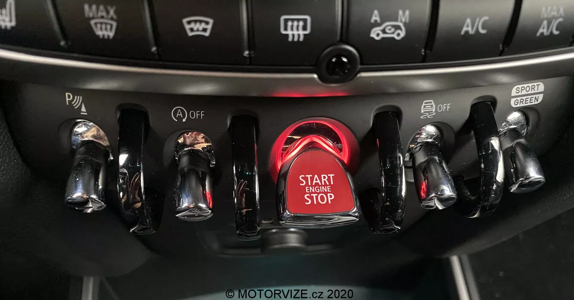 Vista aproximada do console central do Mini Countryman John Cooper Works 2020, focando na área de ignição com o botão de start/stop do motor em destaque e cercado por um anel vermelho brilhante, com interruptores metálicos e acabamento cromado em ambos os lados para diversos controles do veículo, incluindo modos de condução e configurações de clima.