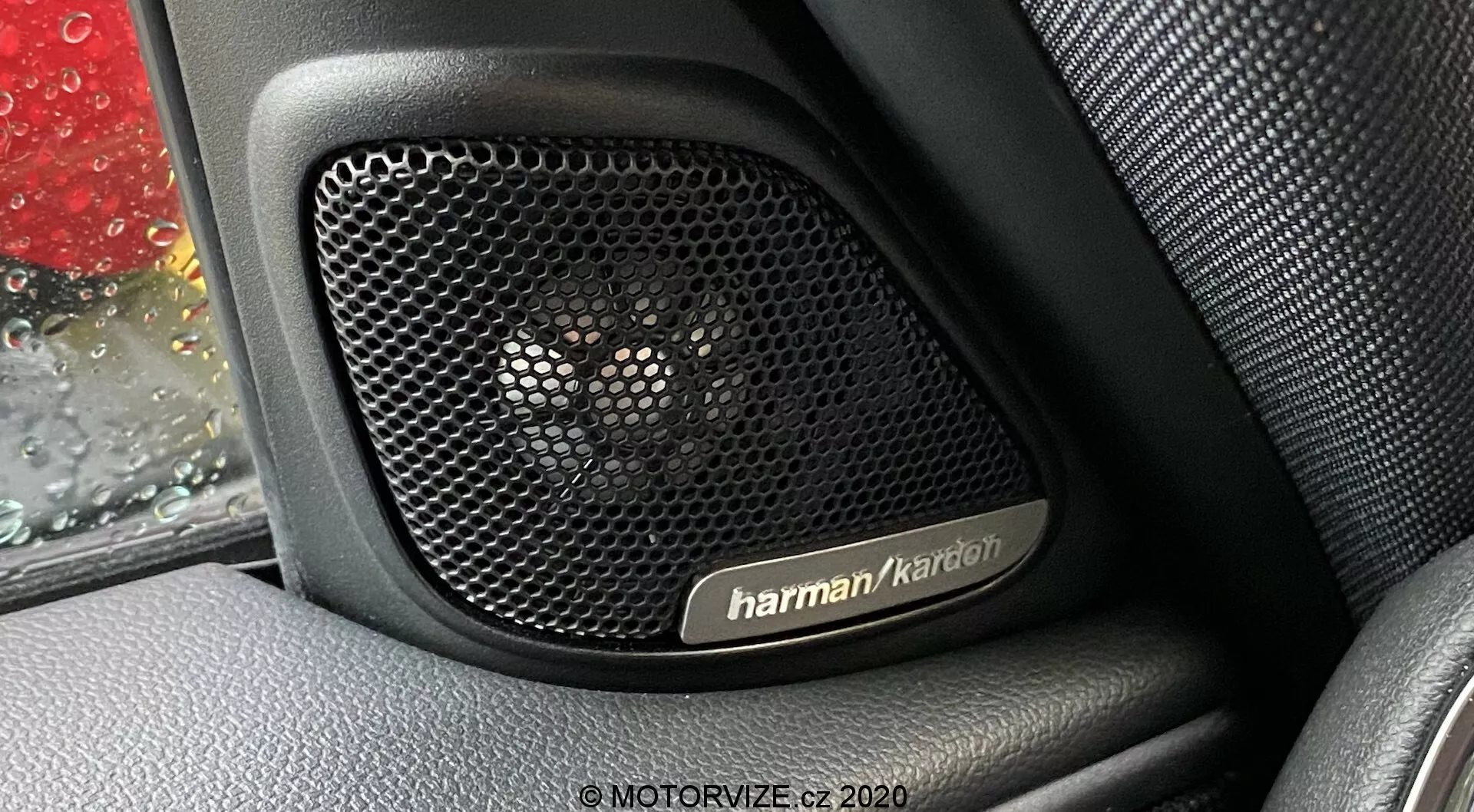 Imagem de close-up do sistema de alto-falantes Harman Kardon no interior da porta de um Mini Countryman 2020 John Cooper Works. A fotografia foca na grade do alto-falante perfurada, projetada para transparência acústica e proteção, contra um interior texturizado parecido com couro. Gotas de chuva são visíveis na janela do carro, adicionando contexto à imagem. O sistema de áudio premium assinala características tecnológicas chave, mostrando o luxo e durabilidade do design do veículo.