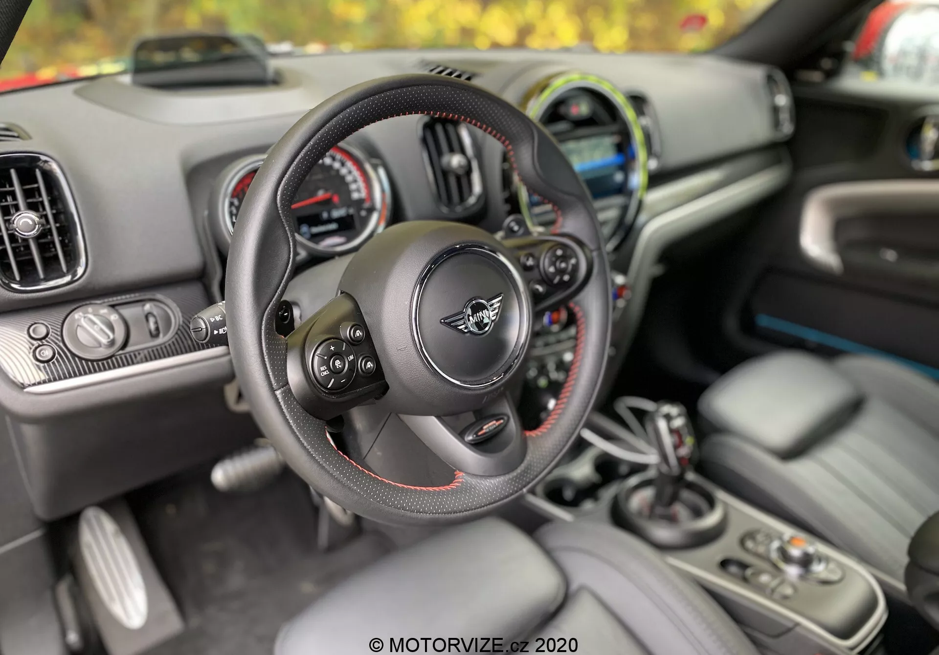 Vista interior do Mini Countryman 2020 na perspectiva do motorista, destacando o volante multifunções com costura vermelha, o painel de instrumentos com mostradores analógicos e digitais, as saídas de ar-condicionado e a tela de infotainment. No console central, são visíveis a alavanca de câmbio, suportes para copos, botões de controle e chave indicando entrada sem chave e botão de partida. O primeiro plano foca no volante e na alavanca de câmbio, destacando a ergonomia e conforto projetados para o motorista.