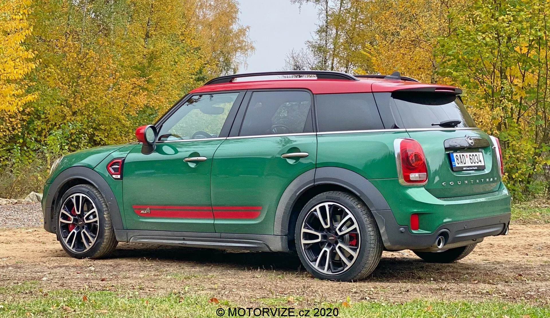 A imagem mostra uma vista traseira de três quartos de um Mini Cooper Countryman verde 2020 com teto e capas de espelhos em vermelho contrastante. Os recursos incluem um spoiler de teto, trilhos de teto, luzes traseiras inspiradas na Union Jack, limpador de para-brisa traseiro, sistema de escape duplo e sensores de estacionamento. O veículo está equipado com grandes rodas de liga e revestimento protetor em torno dos arcos das rodas. A foto foi tirada ao ar livre em uma superfície de cascalho com grama e folhagem ao fundo.