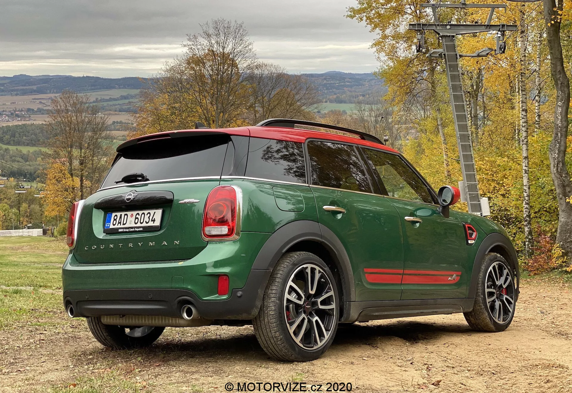 Vista traseira de três quartos de um Mini Cooper Countryman John Cooper Works 2020 verde estacionado ao ar livre, destacando o design do carro da perspectiva lateral traseira. Detalhes notáveis incluem uma carroceria verde escuro com teto vermelho, emblema 'Countryman', logotipo da Mini na tampa traseira, luzes traseiras distintas, sistema de escape duplo, rodas de liga leve, teto panorâmico, limpador do vidro traseiro, janelas traseiras escurecidas, sensores de estacionamento e um retrovisor lateral com indicador de direção integrado. O veículo está em uma superfície não pavimentada com folhagem de outono e uma estrutura de teleférico ao fundo, sugerindo um estilo de vida ativo ao ar livre.