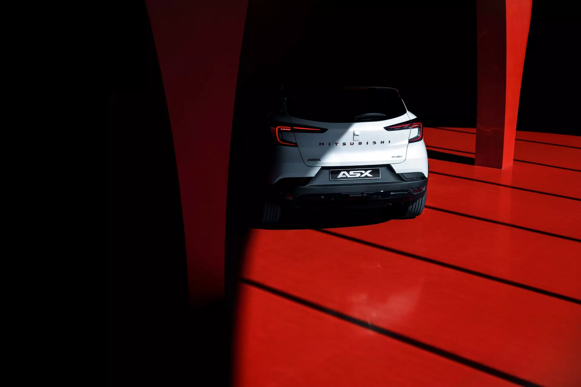 Foto em ângulo baixo da traseira do SUV compacto Mitsubishi ASX 2023 com as luzes traseiras esculpidas, emblema cromado e vidro traseiro, capturado durante o dia com alto contraste em um chão vermelho vivo.