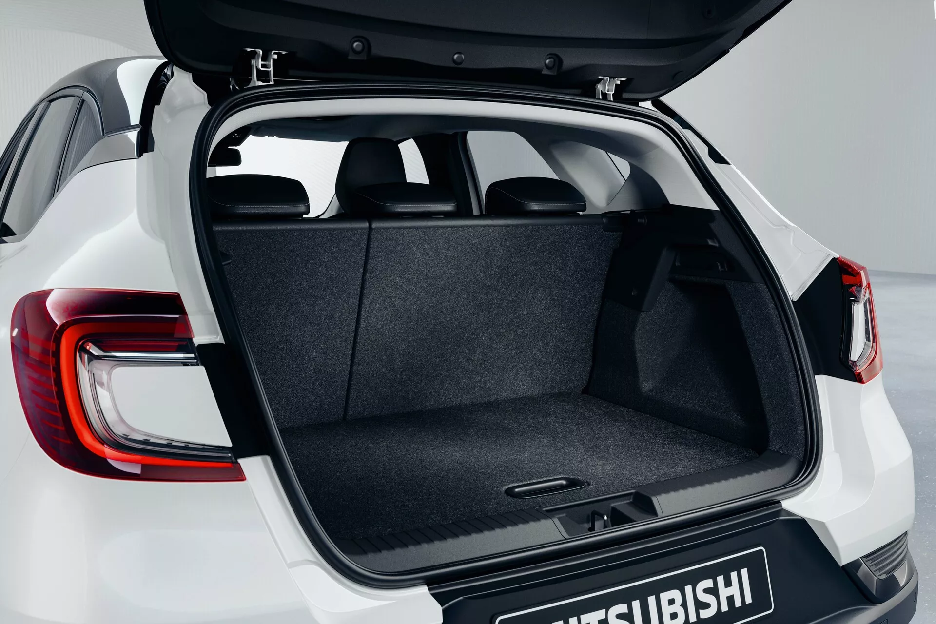Compartimento traseiro do Mitsubishi ASX 2023 (2ª geração) exibindo o porta-malas aberto em um ângulo ligeiramente elevado, destacando o espaço de carga acarpetado, o mecanismo de dobradiça, o piso plano e as luzes traseiras LED modernas parcialmente visíveis com o logotipo da Mitsubishi na porta do porta-malas.