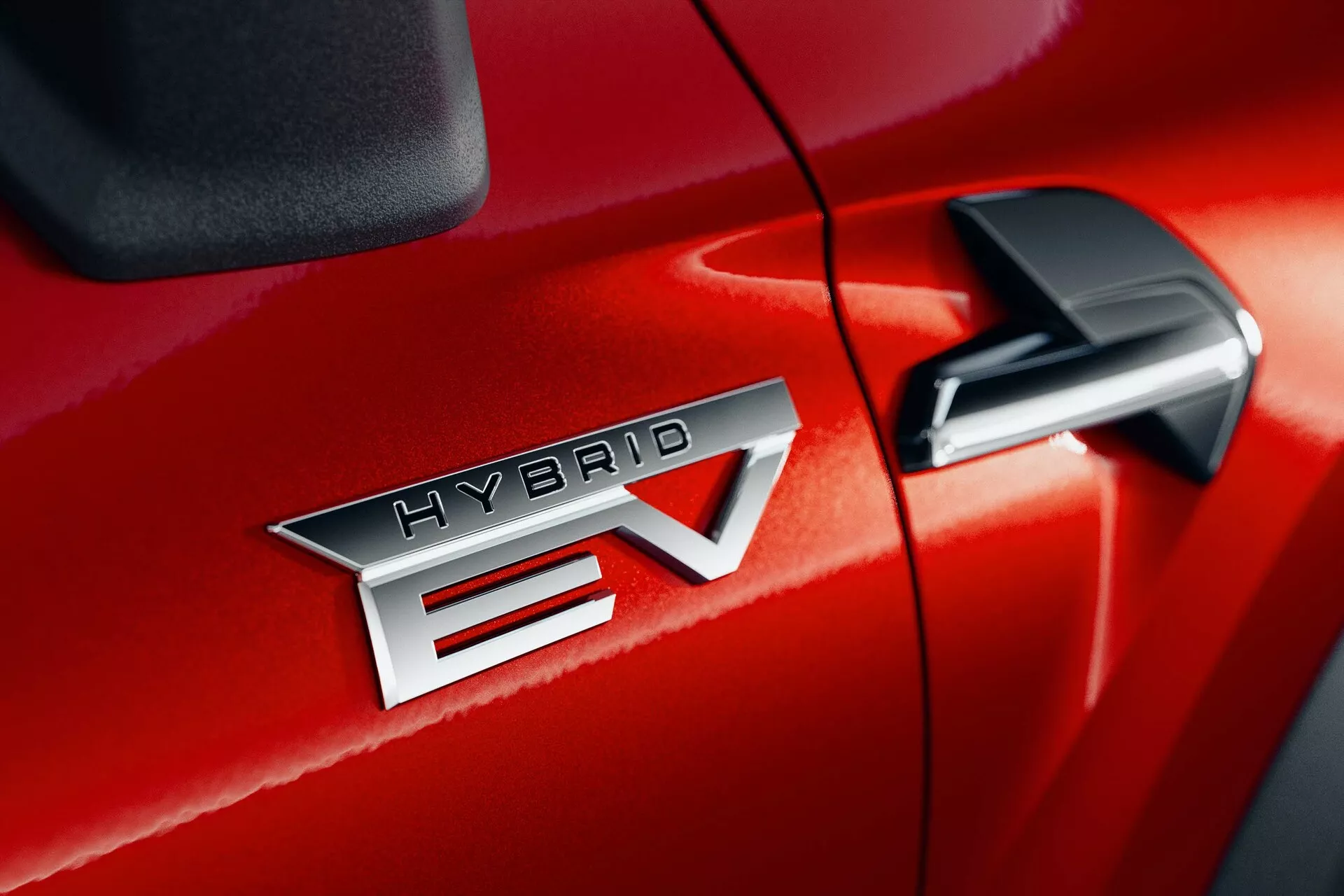 Detalhe em close-up do Mitsubishi ASX 2023 (2ª geração) em vermelho, destacando o emblema 'HYBRID EV' que indica a tecnologia de veículo elétrico híbrido. O emblema 'HYBRID EV' está centrado, destacando o acabamento metálico e o letreiro preciso. O 'EV' apresenta um design distintivo com uma seta através do 'V'. O espelho retrovisor lateral é parcialmente visível no canto superior direito, sugerindo características tecnológicas adicionais. O acabamento brilhante da pintura e a costura apertada entre a porta do passageiro dianteiro e o para-lama dianteiro sugerem uma qualidade superior na construção.