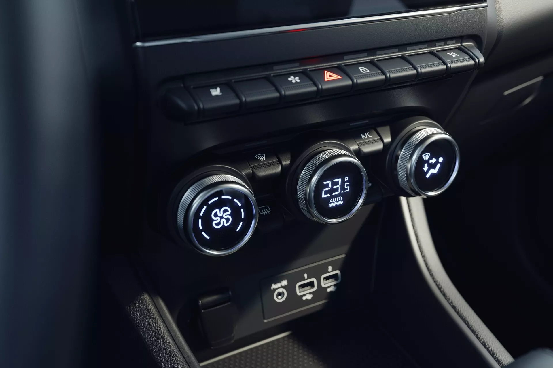 Vista aproximada do sistema de controle de climatização interior do Mitsubishi ASX 2023 (2ª geração). A imagem, tirada de um ângulo elevado, foca na consola central com três dials de controle de temperatura - o esquerdo ajustado para 'LO' e o direito para '23.5'. O dial central parece controlar a velocidade ou direção do ventilador. Acima desses dials, há botões para desembaçar, luzes de emergência e circulação de ar. Abaixo estão portas USB modernas. O design é minimalista com superfícies negras elegantes.