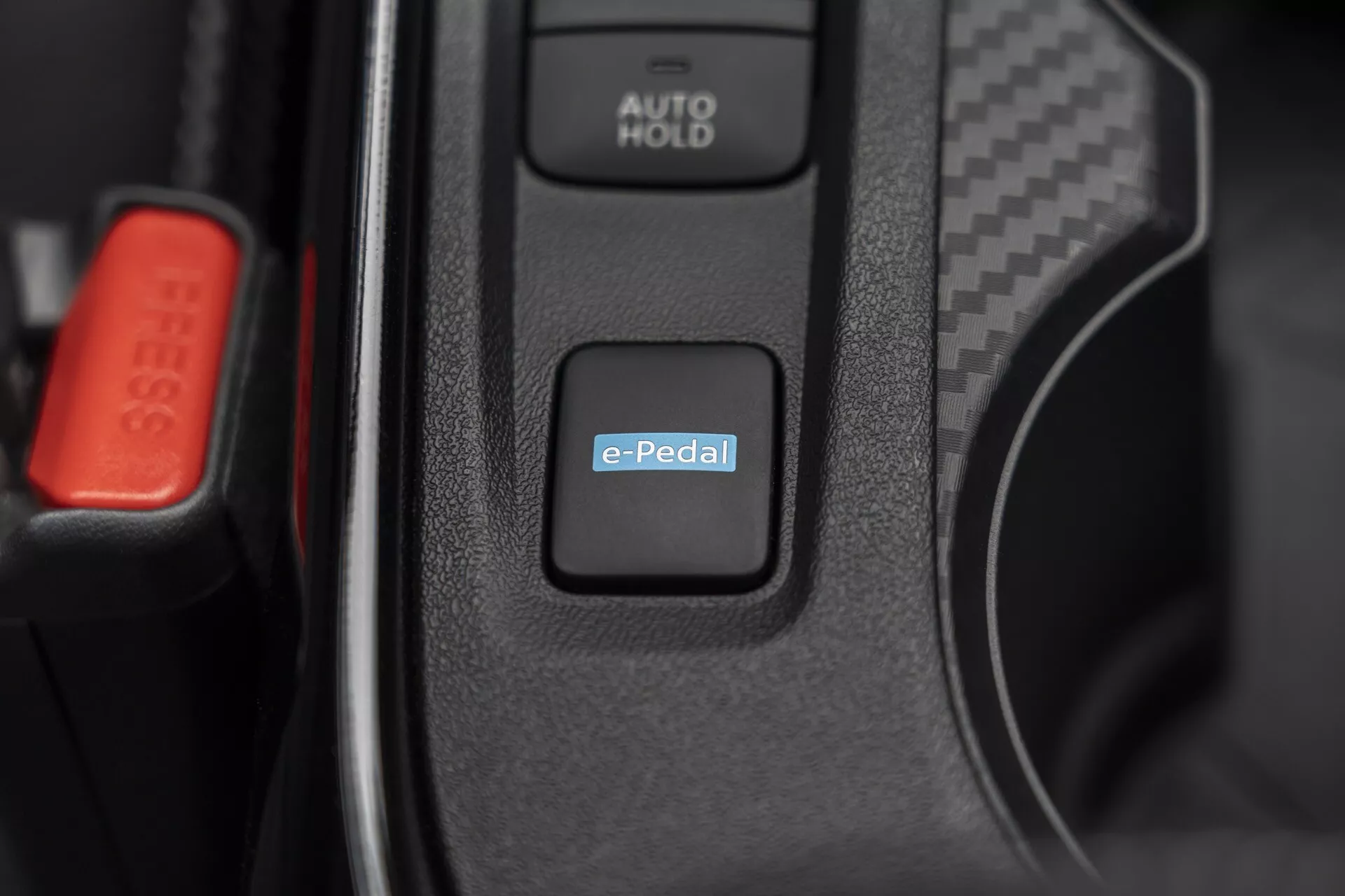 Close-up do interior do Nissan Juke (F16) 2019 destacando o interruptor do sistema e-Pedal para controle de condução elétrica ou híbrida, com botão AUTO HOLD e clip de cinto de segurança vermelho visíveis sugerindo tecnologia automotiva avançada e características interiores premium