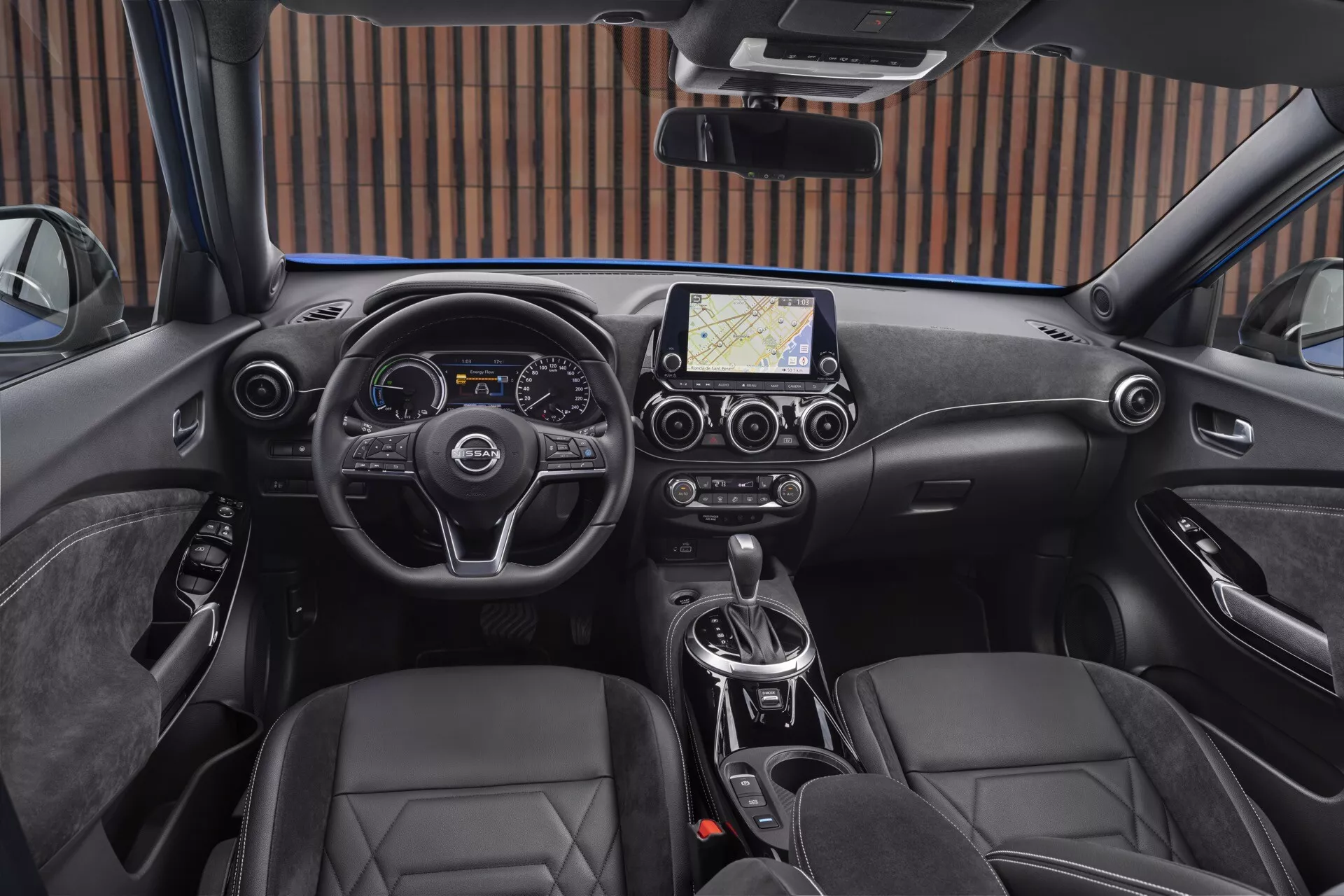 Vista interior do Nissan Juke F16 de 2019 a partir da perspectiva do motorista, apresentando um volante moderno de três raios com botões de controle, cluster de instrumentos digital e analógico combinados, sistema de infotainment com tela sensível ao toque central e navegação, controle climático de duas zonas, transmissão automática, assentos tipo balde de couro e freio de estacionamento eletrônico.