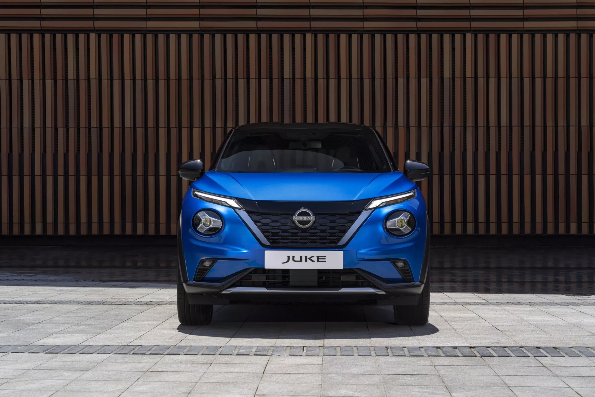 Vista frontal de um Nissan Juke F16 de 2019 em azul, exibindo faróis LED elegantes, grade V-motion, e para-choque esportivo contra um fundo de parede com linhas horizontais