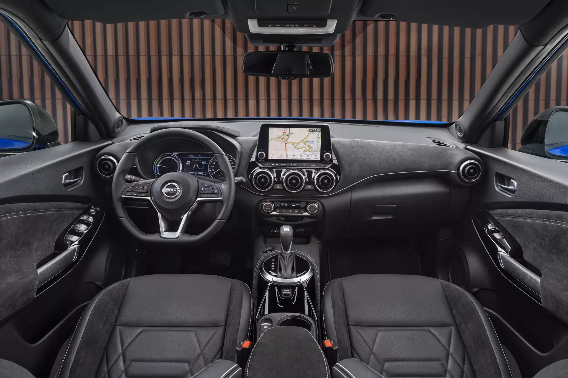 Vista interior do Nissan Juke F16 2019 a partir do banco traseiro em direção ao painel de instrumentos mostrando o volante multifuncional, mostradores dos instrumentos, tela de infotainment com navegação GPS, controle climático, alavanca de câmbio automático, assentos dianteiros estilo esporte com revestimento em couro e recursos de segurança