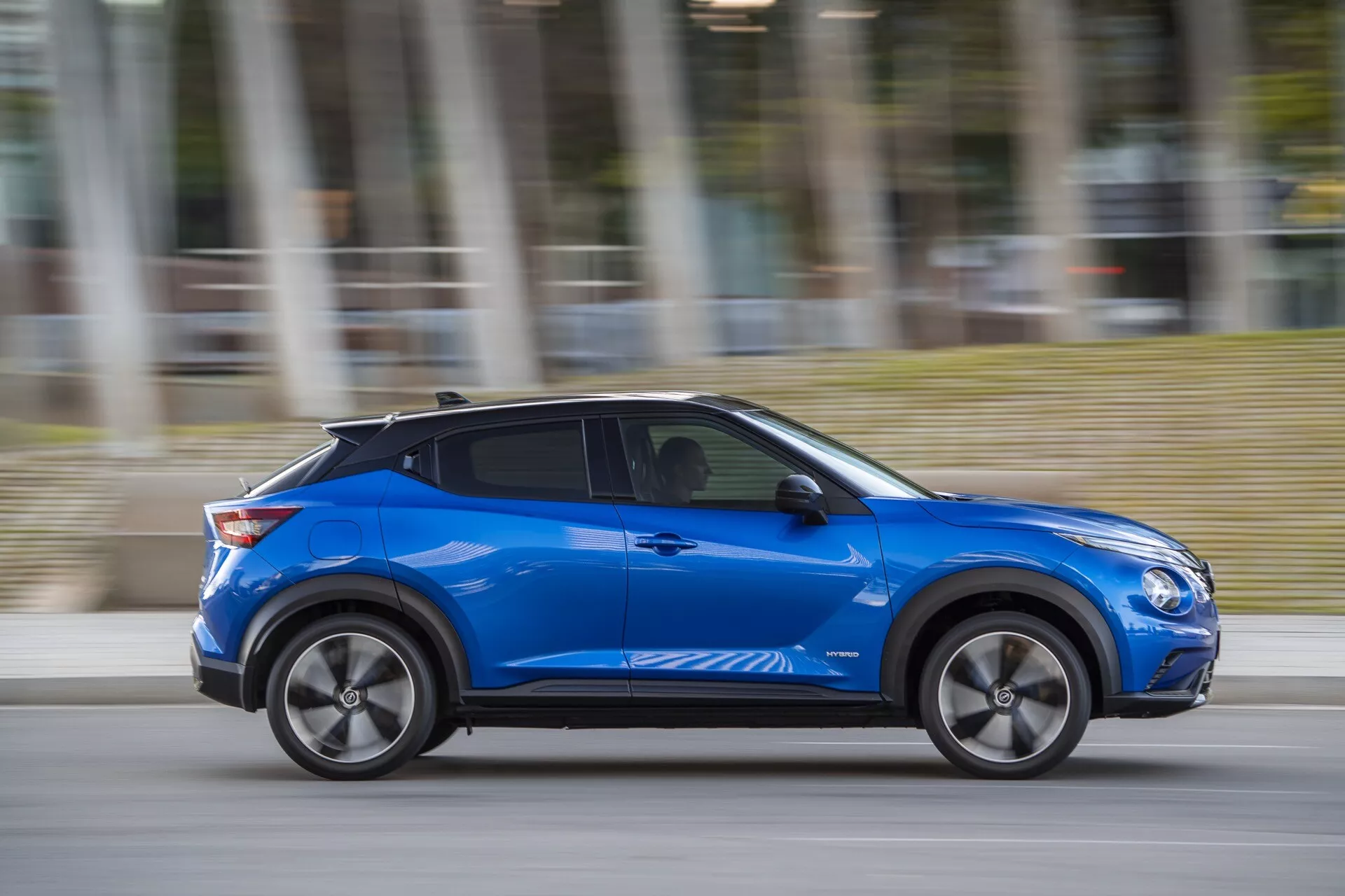 Nissan Juke (F16) 2019 em azul, exibindo design moderno e características impulsionadas pela tecnologia como linhas de corpo aerodinâmicas, rodas de liga leve e entrada sem chave potencial, movendo-se dinamicamente com desfoque de movimento em um dia nublado