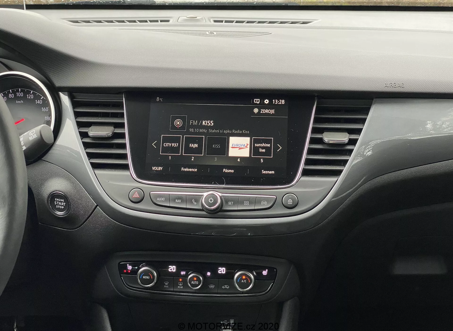 Vista interna do painel do Opel Crossland 2021, mostrando um sistema de infotainment com tela sensível ao toque central exibindo informações de estação de rádio, com botões e mostradores de controle para ajustes de áudio e clima, saídas de ar elegantes e parte do painel de instrumentos, destacando a tecnologia de veículo moderna e o design interior.