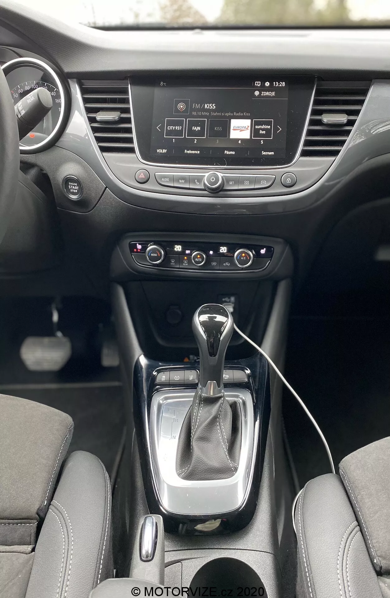 Interior do Opel Crossland 2021 a partir de uma perspectiva de primeira pessoa, mostrando a consola central e o painel de instrumentos. Apresenta uma alavanca de câmbio automático moderna com manga de couro, painel de controle de clima de duas zonas, sistema de infoentretenimento com tela sensível ao toque, porta-copos e volante com controles embutidos.