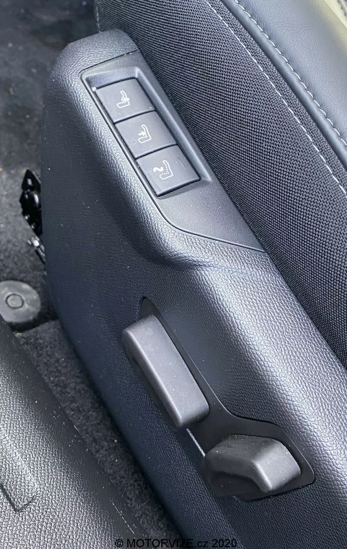 Imagem em close-up dos controles do assento interior do Peugeot 2008 1.2 PureTech 100 Active (2020), destacando o lado de um assento de couro preto ou cinza escuro com costura detalhada, visto de um ângulo oblíquo ligeiramente elevado. O assento apresenta três botões de ajuste eletrônico e uma alavanca manual. Os botões controlam o movimento para frente/para trás, a inclinação do encosto e a altura total do assento, montados em um painel texturizado correspondente. A marca d'água lê-se 'MOTORVIZE.cz 2020'.