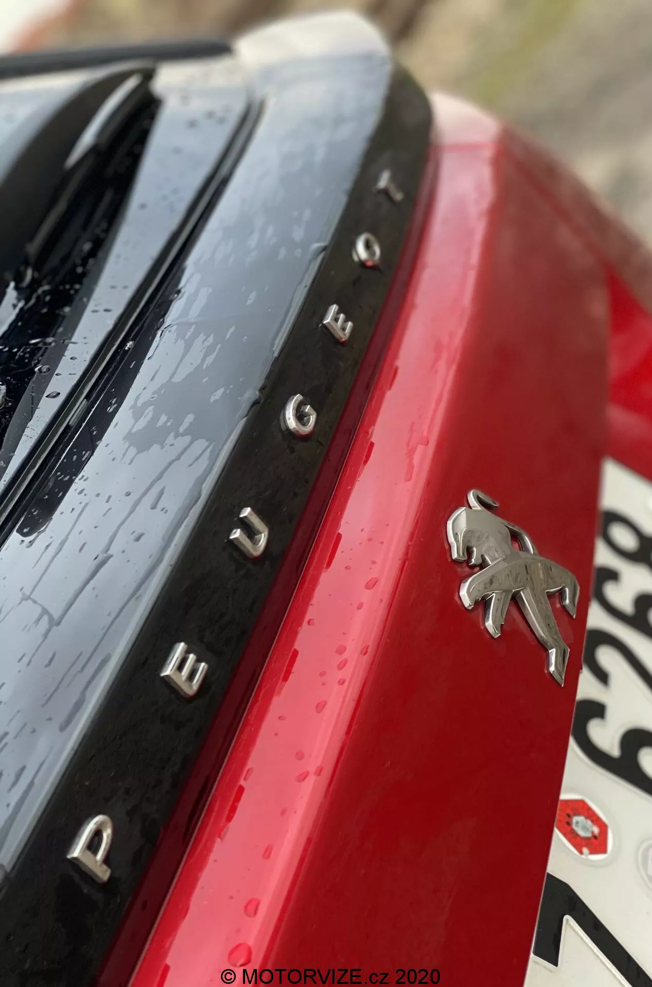 Detalhe em close-up do emblema traseiro e da designação do modelo Peugeot 2008 2020 (1.2 PureTech 100 Active). A foto está em um ângulo diagonal não ortodoxo, capturando o logotipo Peugeot cromado contra o painel de carroceria vermelho vibrante. Letras cromadas espaçadas à esquerda do logotipo formam 'PEUGEOT'. Uma superfície preta brilhante com gotas de água aparece acima do nome, indicando chuva recente ou uma lavagem de carro. A seção da placa desfocada é visível abaixo e à direita. A pequena profundidade de campo foca nitidamente o emblema e o nome, enquanto os outros elementos estão desfocados para um toque artístico.