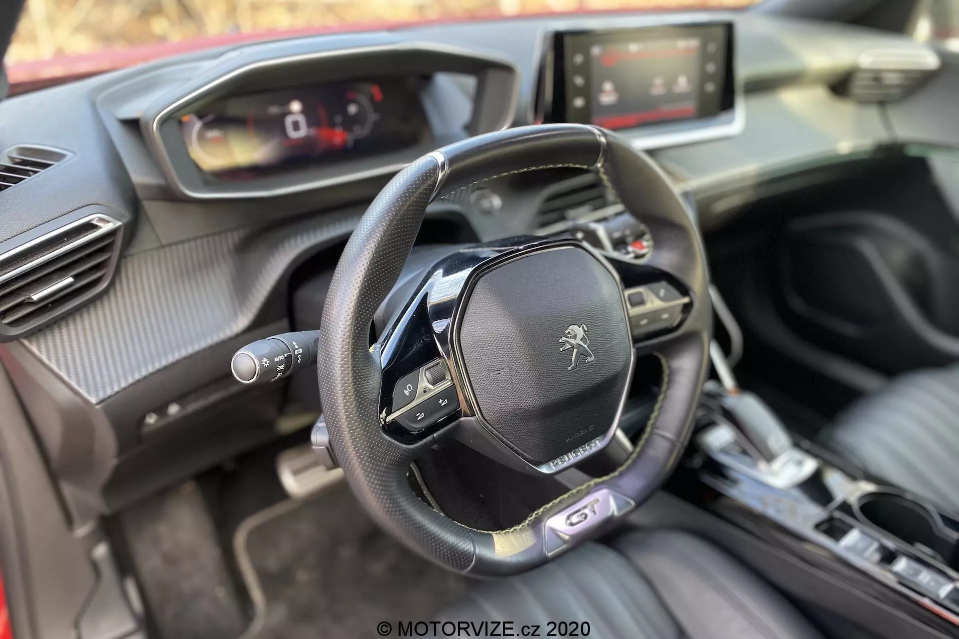 Interior do Peugeot 2008 1.2 PureTech 100 Active de 2020 a partir da perspectiva do passageiro da frente focado no cockpit do motorista. A imagem mostra um volante de fundo plano com vários botões de controle e o logotipo do leão da Peugeot, um painel de instrumentos digital, sistema de infoentretenimento com tela sensível ao toque, console central de transmissão automática, botão de ignição start/stop, saídas de ar com detalhes cromados e uma mistura de materiais macios ao toque, incluindo couro e texturas semelhantes à fibra de carbono.