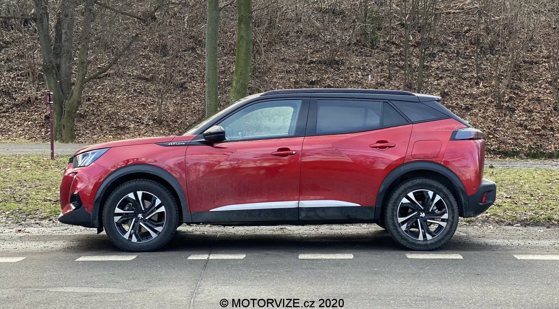 Veículo utilitário esportivo (SUV) Peugeot 2008 2020 1.2 PureTech 100 Active vermelho com design moderno e elegante, linha do teto inclinada, linha de cintura alta com janelas afuniladas, maçanetas aerodinâmicas, espelhos laterais com luzes indicadoras integradas, grandes rodas de liga de múltiplos raios, pneus de perfil baixo, revestimentos de plástico preto para um visual robusto, pintura vermelha brilhante e vidros traseiros escurecidos, capturado em vista de perfil estacionado em um ambiente ao ar livre com árvores sem folhas e fundo de grama.
