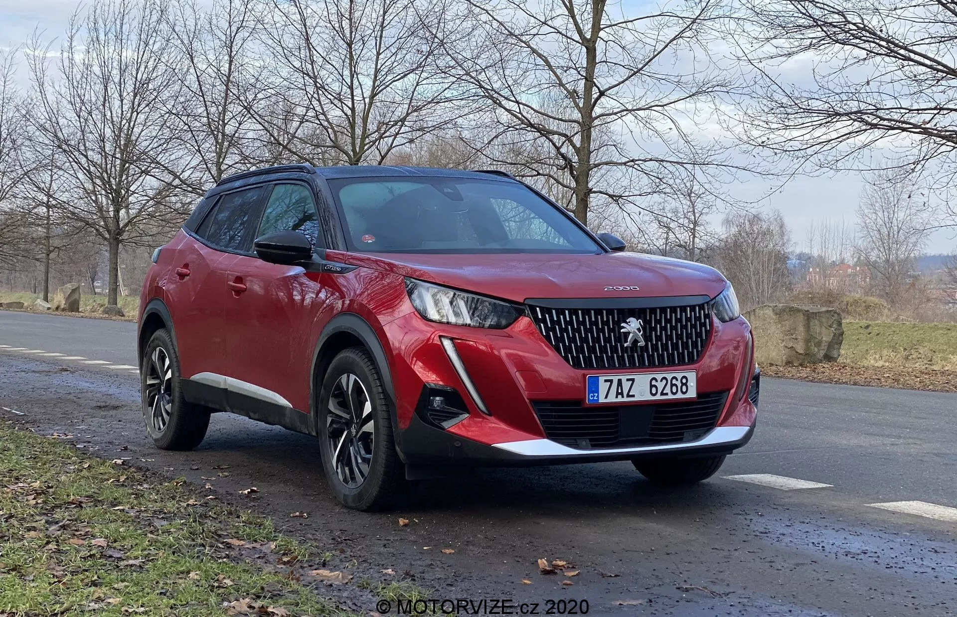 SUV vermelho Peugeot 2008 2020 (Peugeot-2008-1-2-Puretech-100-Active) da vista frontal de três quartos, destacando a grade frontal com um emblema de marca proeminente, faróis LED elegantes, sensores de estacionamento dianteiros e luzes de neblina. A vista lateral revela rodas de liga leve de vários raios, maior altura de direção, linhas dinâmicas da carroceria, sinais de direção integrados nos espelhos laterais e revestimento de plástico preto. Posicionado em ângulo leve ao lado da estrada com paisagem rural natural ao fundo, indicando final de outono ou início da primavera. Ângulo de câmera baixo para uma vista poderosa e imponente.