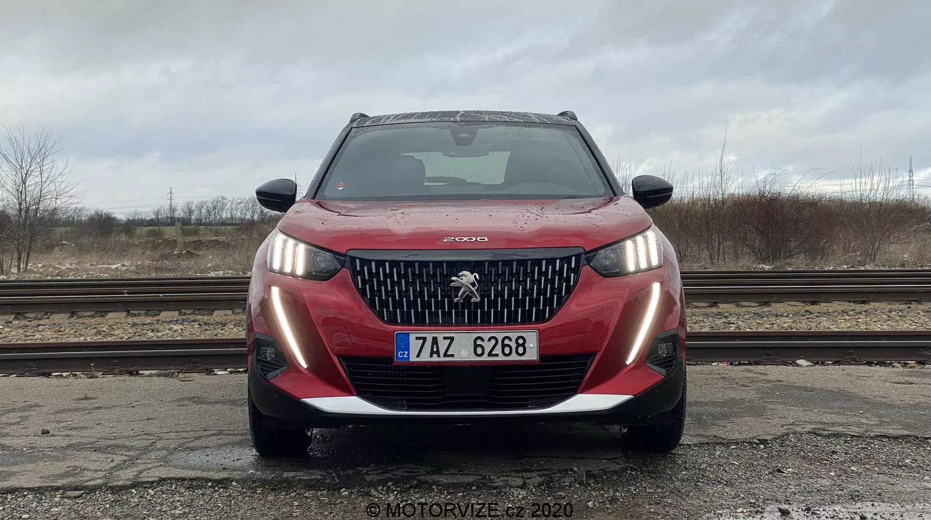 Vista frontal de um modelo Peugeot 2008 2020 moderno, capturado de uma perspectiva de ângulo baixo. A imagem mostra a grade frontal do carro, o logo distintivo do leão, faróis de LED e o para-choque. A grade apresenta lâminas verticais típicas da linguagem de design da Peugeot. O ângulo enfatiza a largura e a presença na estrada do veículo. O para-choque inclui elementos na cor do veículo e em preto, sugerindo um design esportivo, com sensores de estacionamento visíveis e uma placa de matrícula no centro. A cena foi tirada em um dia nublado com um fundo desfocado e árvores nuas à distância.