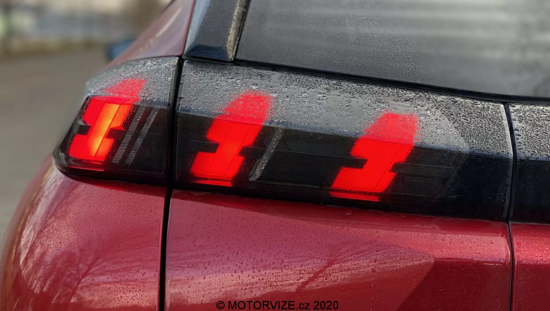 Close-up da seção traseira de um Peugeot 2008 2020 vermelho (PureTech 100 Active) com foco na montagem da lanterna traseira mostrando as luzes de freio acesas. A fotografia, tirada do canto traseiro ligeiramente de lado, destaca três elementos de luz LED verticais distintos e gotas de água na superfície, enfatizando o design moderno da tecnologia de iluminação automotiva.