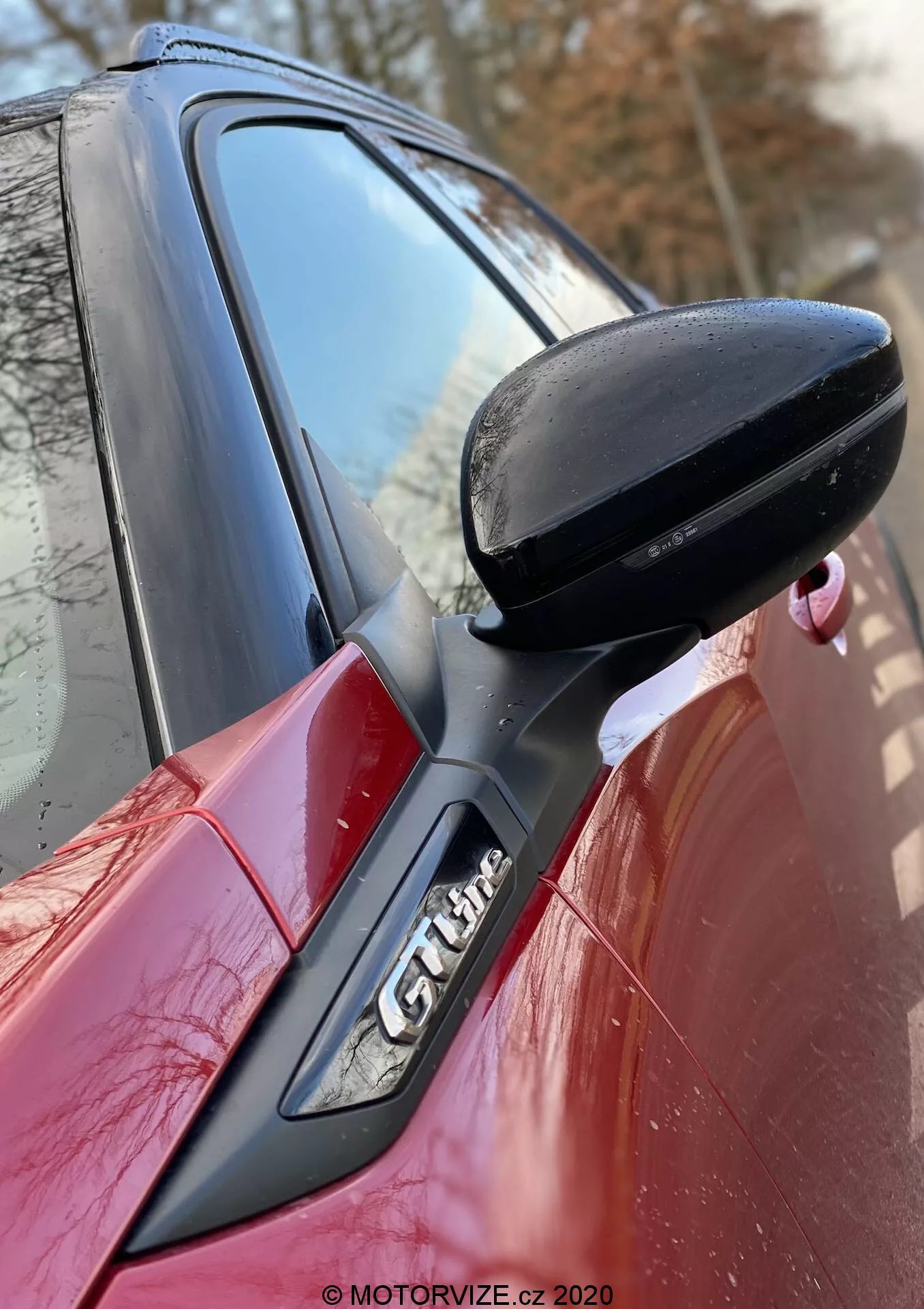 Close-up da lateral de um Peugeot 2008 2020 vermelho, focando no espelho retrovisor do lado do motorista preto, montado na porta com suporte preto. O pilar A com o emblema 'GT Line' é parcialmente visível, mostrando as linhas de design aerodinâmico do carro e as características esportivas. Marca d'água da MOTORVIZE.cz 2020.