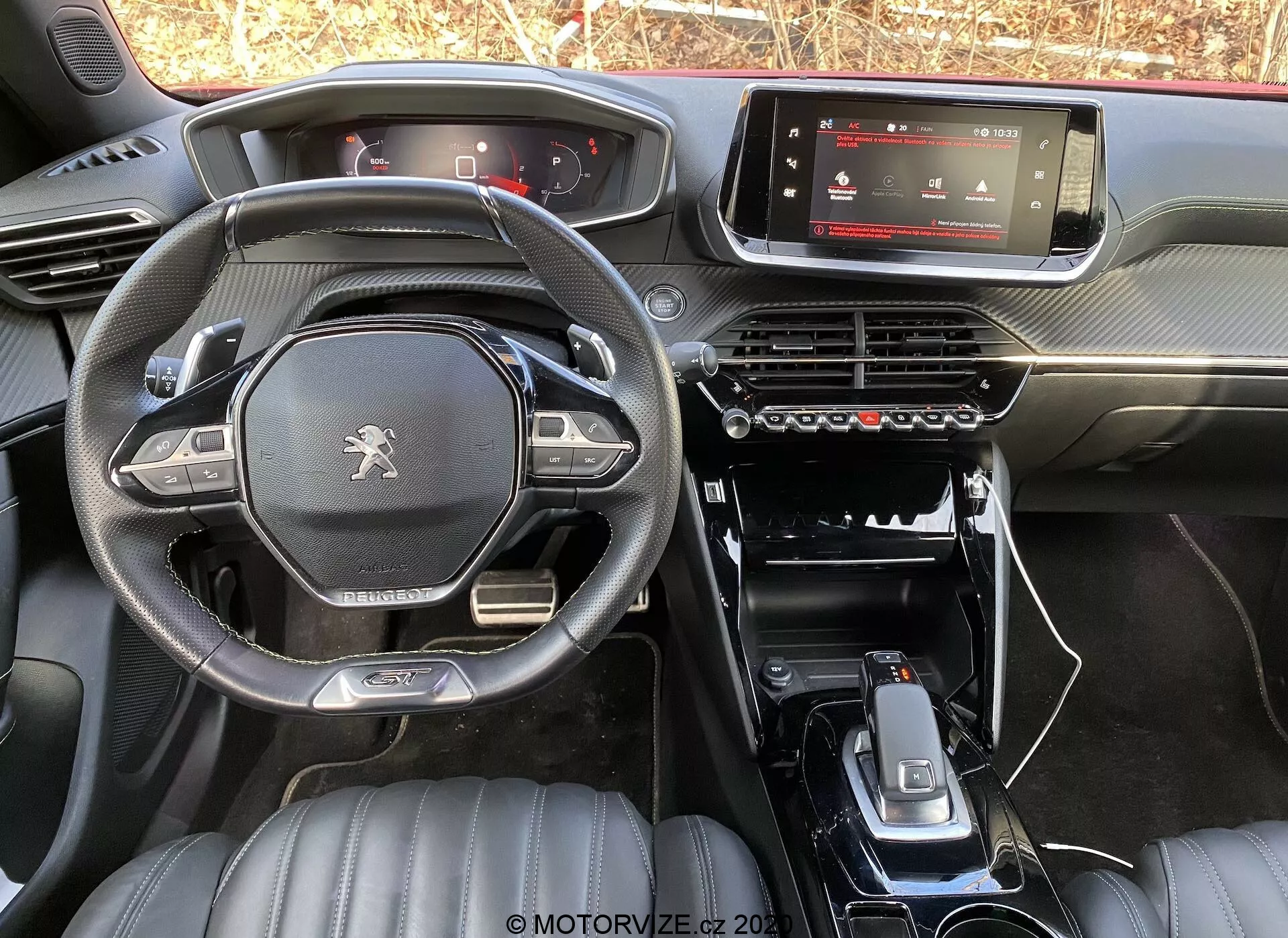 Vista interior do Peugeot 2008 1.2 PureTech 100 Active 2020 a partir do banco traseiro, mostrando o painel de instrumentos, tela digital de infotainment, painel de controle de climatização, console central minimalista com alavanca de câmbio, suportes para copos e botões de controle adicionais. O volante de fundo plano apresenta o logotipo Peugeot e controles multifuncionais. Os assentos dianteiros em couro com padrões acolchoados sugerem um nível de acabamento superior. A iluminação natural destaca o design e os materiais do modelo de fábrica.