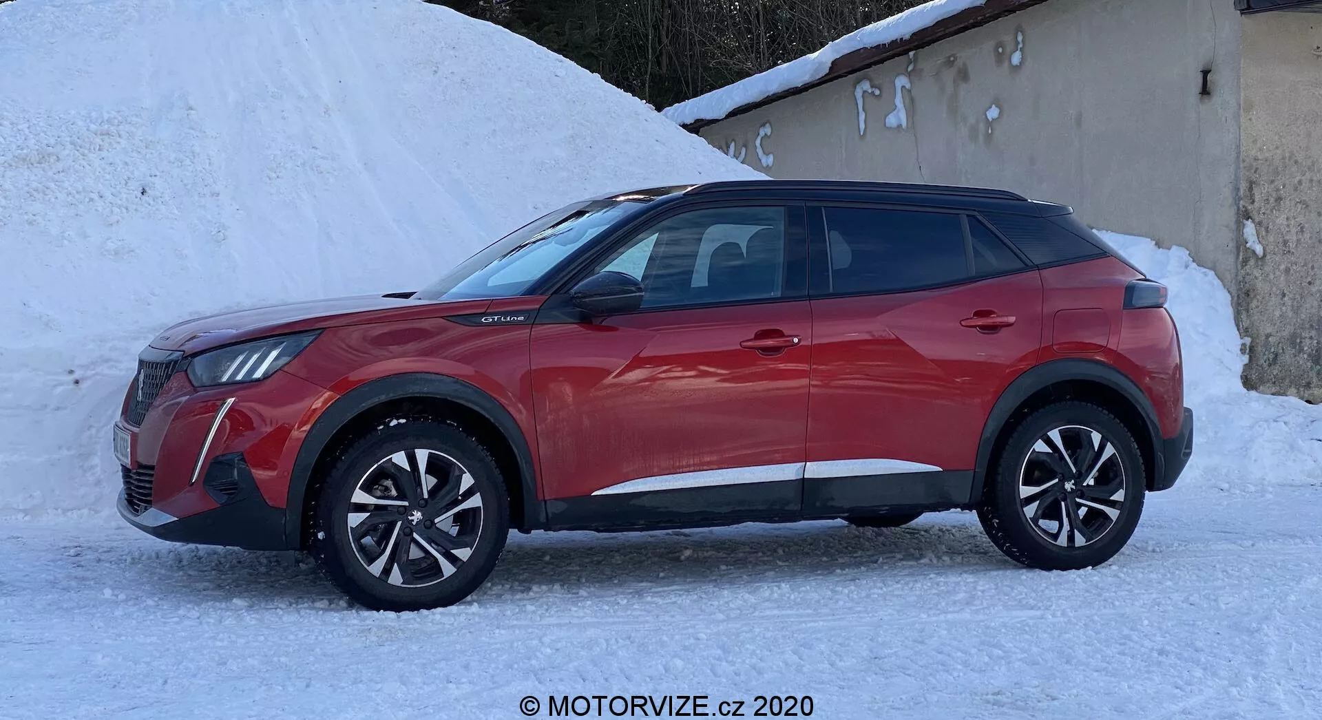 Foto de perfil lateral de um SUV crossover Peugeot 2008 2020 1.2 PureTech 100 Active de quatro portas vermelho. O veículo está em um ambiente nevado, enfatizando suas potenciais capacidades em clima frio. Possui rodas de liga leve com design de raios múltiplos, revestimento de plástico preto ao longo da parte inferior das portas e dos arcos das rodas, e janelas elegantes. O design dos faróis e a grade são visíveis na parte frontal, mostrando uma aparência moderna com prováveis ​​faróis de LED. Os retrovisores laterais incluem sinais de seta integrados. A linha do teto inclinada e o sistema de trilhos do teto sugerem praticidade e utilidade para um estilo de vida ativo.