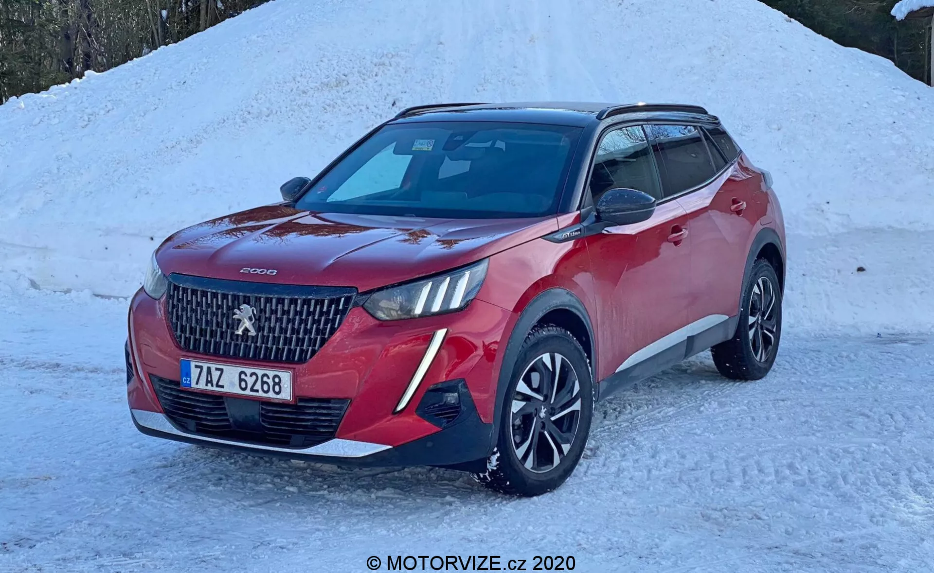 Imagem de um SUV vermelho, especificamente um Peugeot 2008 2020 (peugeot-2008-1-2-puretech-100-active), estacionado em um terreno coberto de neve. O veículo moderno é mostrado em um ângulo de três quartos da frente, destacando sua grade proeminente com um grande logotipo central e design de padrão de diamante. Faróis elegantes em LED, pintura de carroceria em duas cores com capas de espelho pretas e teto, rodas de liga de design multiraio e faróis de neblina ou iluminação adicional no para-choque dianteiro são visíveis. O carro possui sensores de estacionamento dianteiros, um módulo de câmera ou sensor no para-brisa para tecnologia de assistência ao motorista e uma antena em formato de barbatana de tubarão no teto, sugerindo funcionalidade de GPS ou rádio via satélite. A cena indica um ambiente de inverno com uma luz natural brilhante que realça os detalhes do carro.