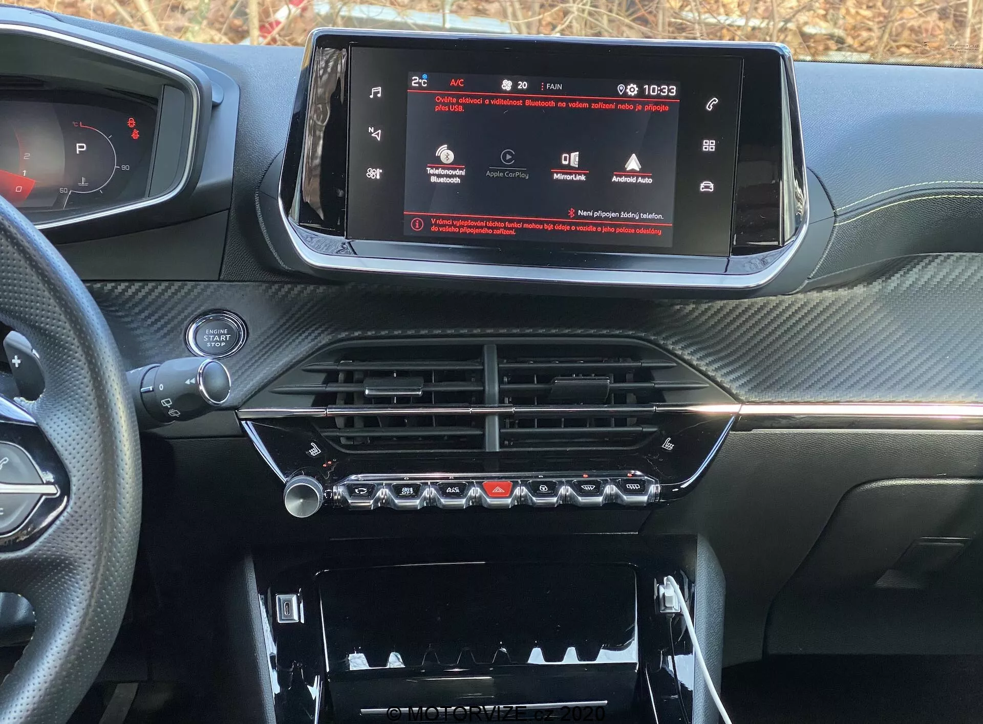 Close-up do painel interno de um carro moderno, especificamente o Peugeot 2008 2020 (peugeot-2008-1-2-puretech-100-active). A imagem é tirada do assento do motorista ou do passageiro dianteiro, focando na console central e no sistema de infoentretenimento. Uma tela widescreen touchscreen mostra um menu de configurações do veículo, indicando tecnologia avançada. Abaixo estão saídas de ar cromadas e controles de clima com botões físicos e botões rotativos. Mais abaixo, há um compartimento com uma tampa visível. O painel tem um acabamento texturizado semelhante ao de fibra de carbono, conferindo um aspecto esportivo e sofisticado.