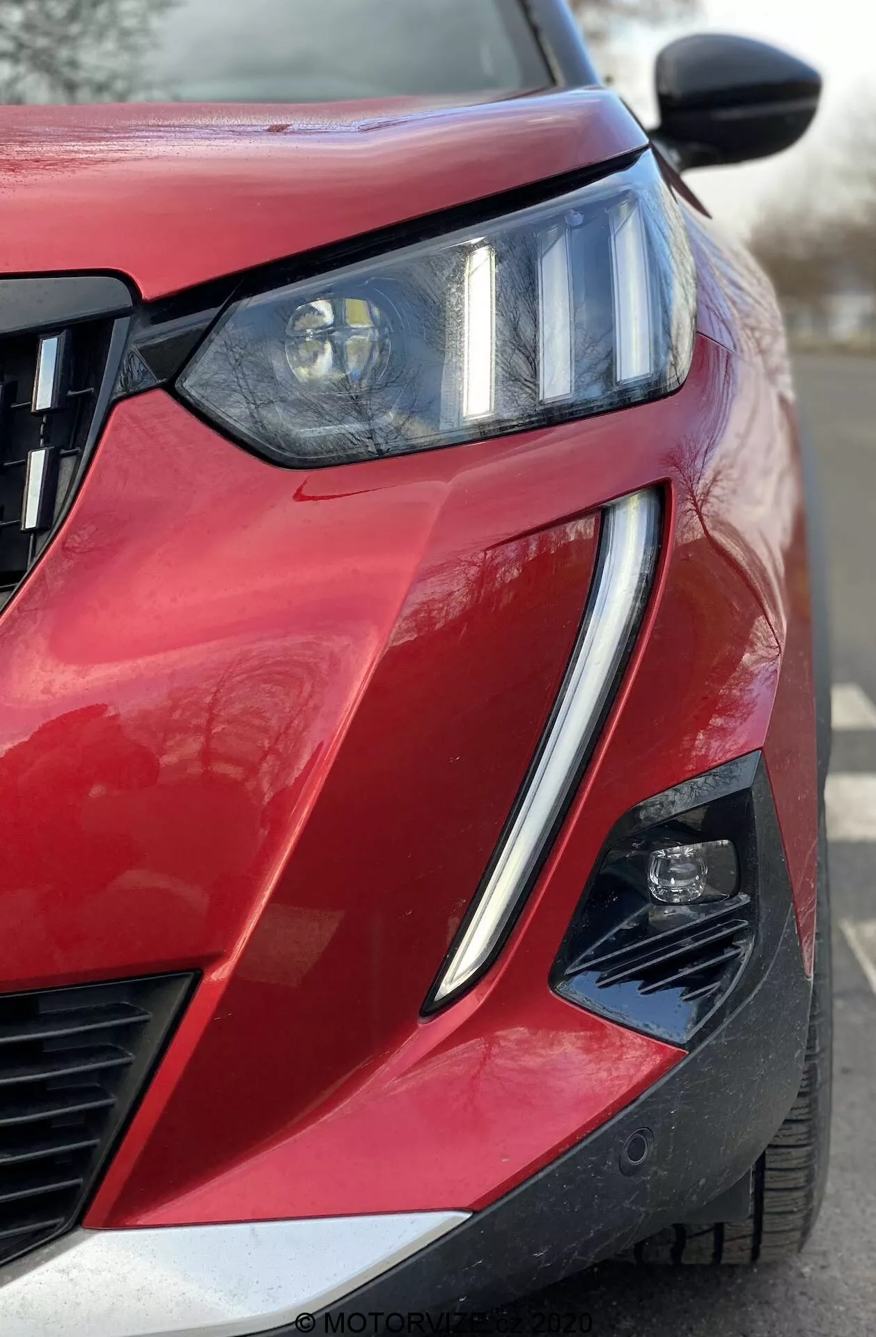 Close-up angular frontal de um Peugeot 2008 1.2 PureTech 100 Active vermelho de 2020, mostrando o quarto dianteiro do lado do motorista. Farol proeminente com feixe de projetor e DRLs de LED integrados, grade de lâminas horizontais e conjunto de luz secundária parcialmente visível dentro de um recorte angular no para-choque. Pintura brilhante com acabamento metálico, linha de carroçaria nítida, contorno elevado do capô, espelho retrovisor lateral preto possivelmente com sinais de mudança integrados ou câmera de ponto cego, e recorte no canto inferior do para-choque sugerindo sistemas avançados de assistência ao motorista.