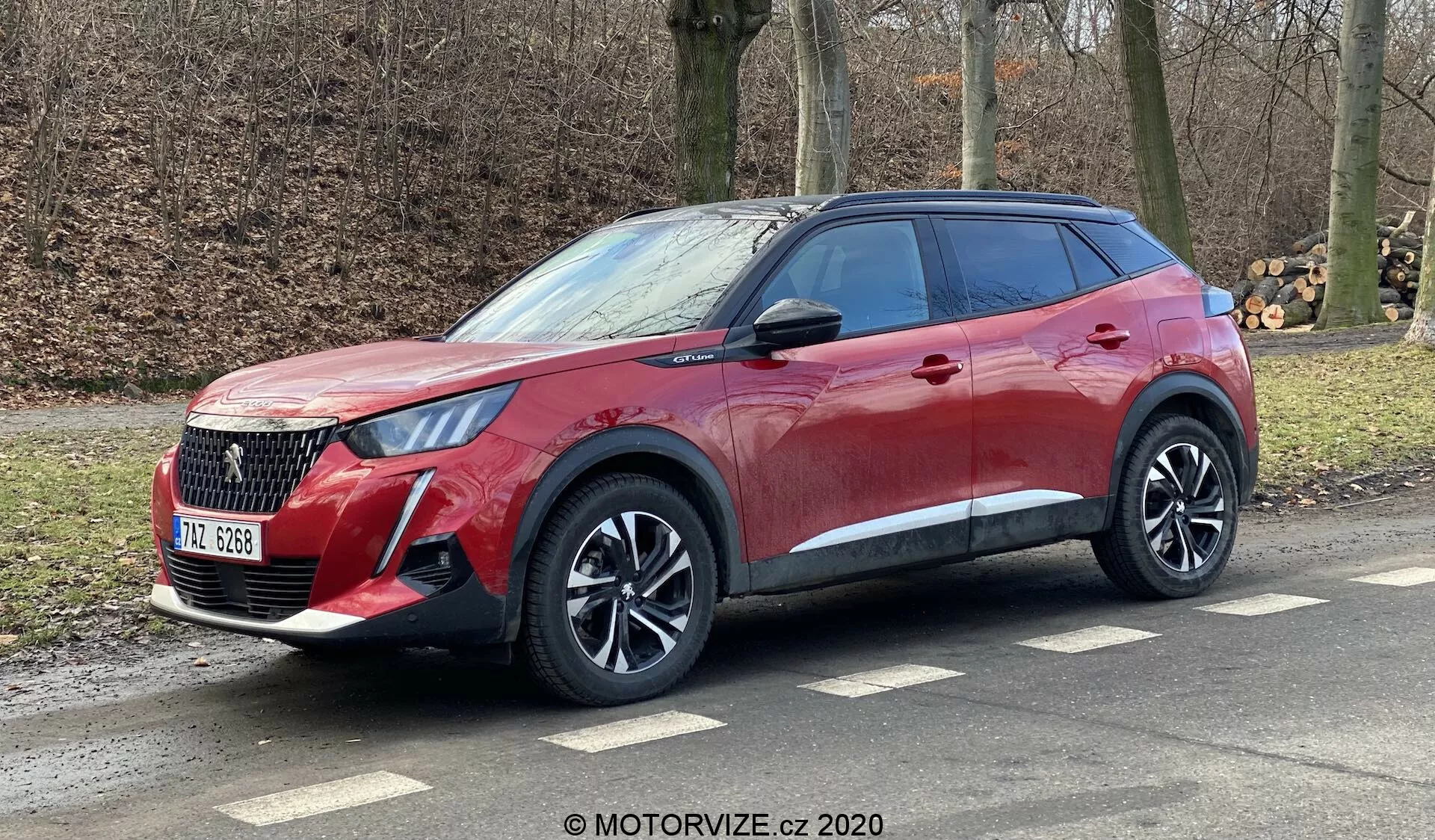 A imagem mostra um Peugeot 2008 1.2 PureTech 100 Active 2020, um SUV moderno estacionado em ângulo na beira da estrada. Esta perspectiva tridimensional destaca a frente e o lado do passageiro do veículo, revelando o acabamento de pintura vermelha elegante, as linhas dinâmicas da carroceria, a grade distinta, os faróis LED nítidos e as rodas de liga leve de múltiplos raios. Os arcos das rodas pronunciados, a linha de cintura ascendente e os detalhes cromados sob as janelas escurecidas aumentam a aparência premium. A linha do teto inclina-se suavemente com barras de tejadilho visíveis, sugerindo versatilidade esportiva. A parte traseira parcialmente visível sugere luzes traseiras envolventes em LED e um spoiler integrado.
