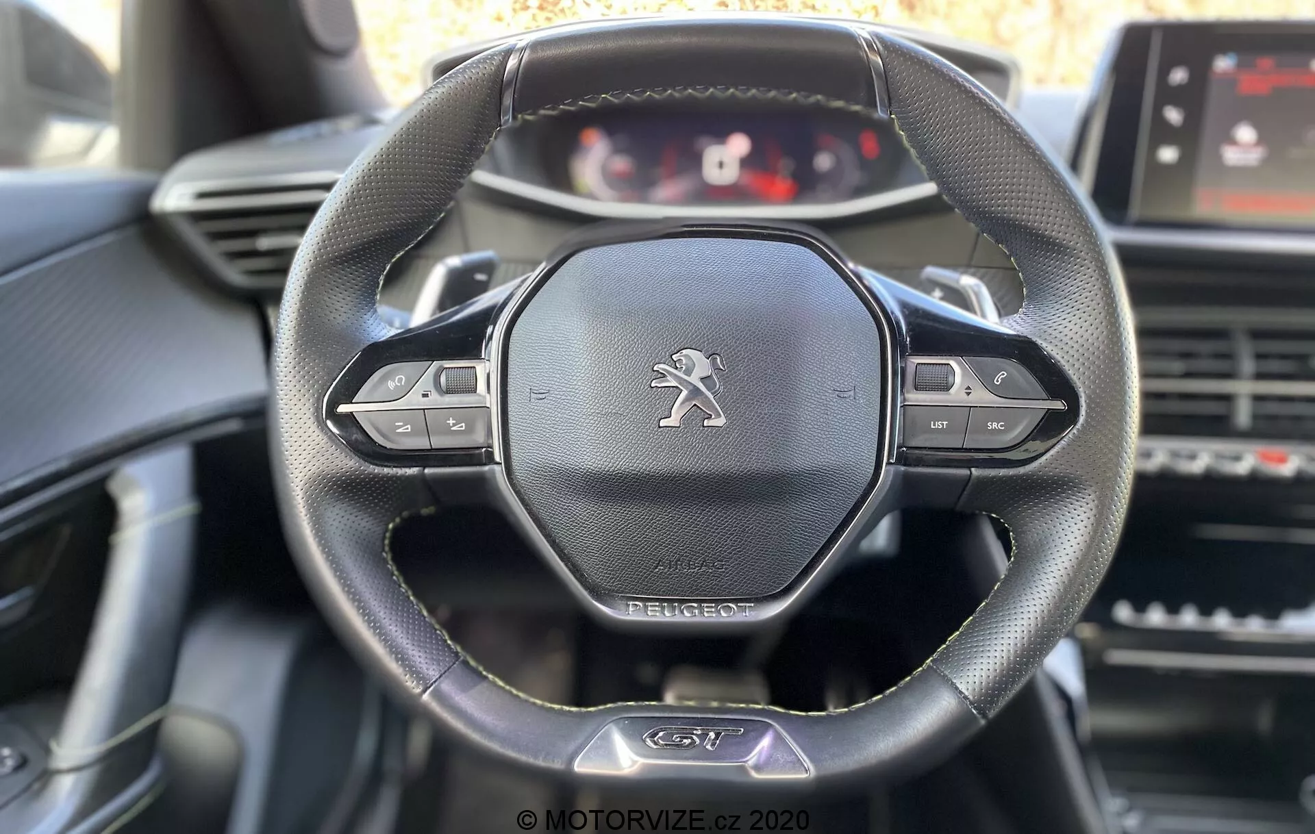 Vista interior do Peugeot 2008 2020 focado no volante e painel de instrumentos da perspectiva do motorista. Recursos principais incluem volante de couro de três raios com botões multifuncionais, emblema do leão Peugeot e painel de instrumentos digital personalizável. Elementos visíveis incluem tela sensível ao toque para infotainment, saídas de ar e controles do console central com iluminação suave de luz natural destacando os detalhes do volante.