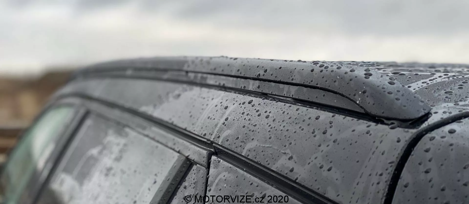 Close-up do teto e da janela lateral do modelo Peugeot 2008 1.2 PureTech 100 Active 2020, destacando gotas de chuva na superfície elegante. A imagem captura a pintura escura e brilhante com gotas de chuva e o design minimalista da borda superior da janela lateral. A pouca profundidade de campo enfatiza a textura exterior do carro enquanto desfoca o fundo e partes do teto, mostrando uma representação artística do carro em condições úmidas.