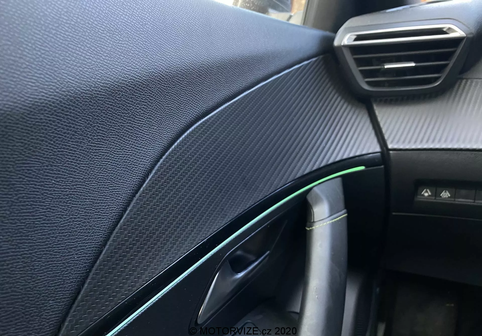 Vista detalhada do painel interior e dos detalhes do lado do passageiro de um Peugeot 2008 2020, focando no painel texturizado preto sintético que remete à fibra de carbono, ventilação ajustável visível, porta parcialmente aberta com reflexo verde e acento cromado, maçaneta da porta, fio com conector de carregamento, iluminação da cabine e um botão não identificado, capturada com pouca profundidade de campo e iluminação natural.