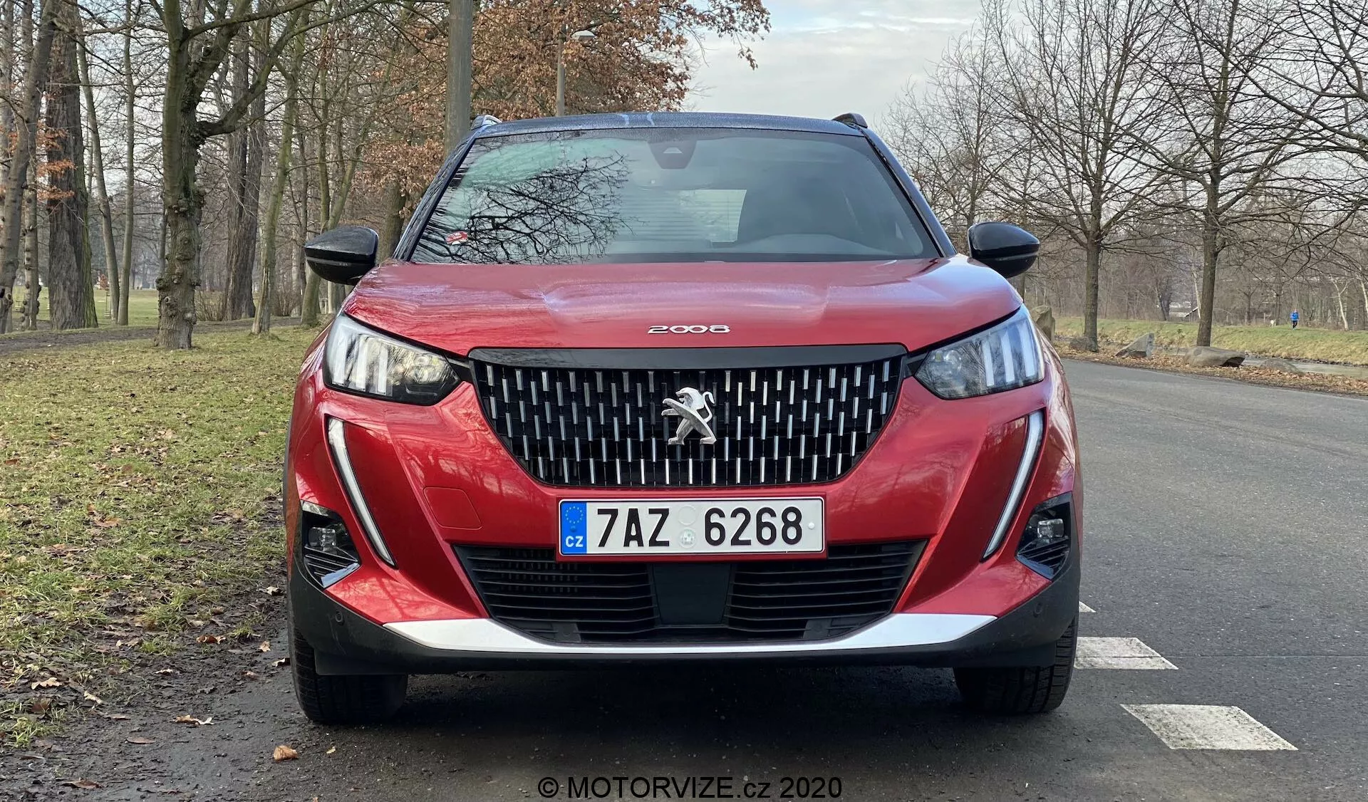 Vista frontal próxima de um Peugeot 2008 2020 vermelho (Peugeot-2008-1-2-PureTech-100-Active) destacando a grade dianteira detalhada com o proeminente emblema do leão Peugeot, design agressivo dos faróis com luzes diurnas de LED em forma de 'dente de sabre', para-choque inferior esculpido e placa de licença checa. O contorno do capô adiciona dinamicidade, e a imagem captura uma visão parcial das rodas de liga leve e espelhos retrovisores laterais em uma estrada de asfalto seco com árvores sem folhas e uma área gramada ao fundo, sugerindo um ambiente natural e sereno, possivelmente no outono ou inverno.