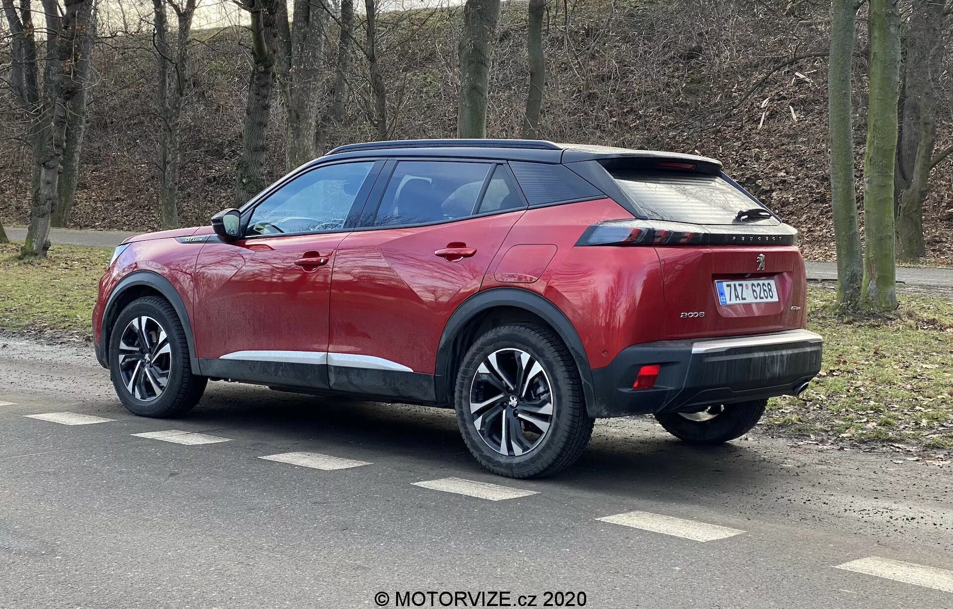 A imagem apresenta um SUV Peugeot 2008 2020 em vermelho metálico brilhante com teto preto contrastante, rodas com múltiplos raios e pneus de perfil baixo. Fotografado em um ângulo traseiro de três quartos, o SUV mostra suas linhas laterais nítidas, spoiler integrado, antena estilo barbatana de tubarão, luzes traseiras horizontais estreitas em LED e um elegante para-choque traseiro com sensores de estacionamento. O veículo possui maçanetas e espelhos laterais na cor da carroceria com piscas integrados, vidros traseiros escurecidos e uma placa de matrícula central abaixo de um possível local para câmera de ré.