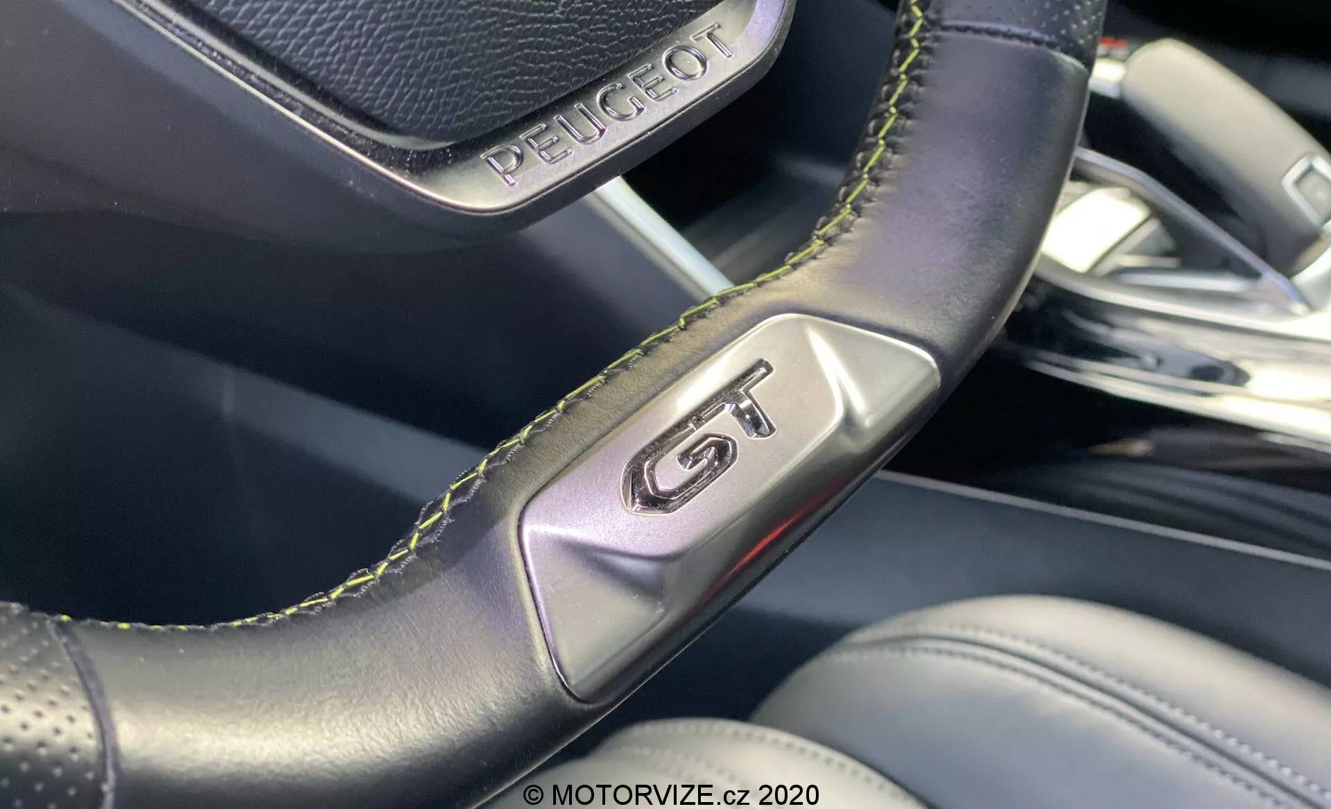 Close-up do interior do Peugeot 2008 2020, destacando o emblema 'GT' no volante de couro perfurado com costura verde, enfatizando esportividade e luxo. A imagem foca nos detalhes artesanais e o fundo desfocado revela materiais internos de alta qualidade e design moderno.
