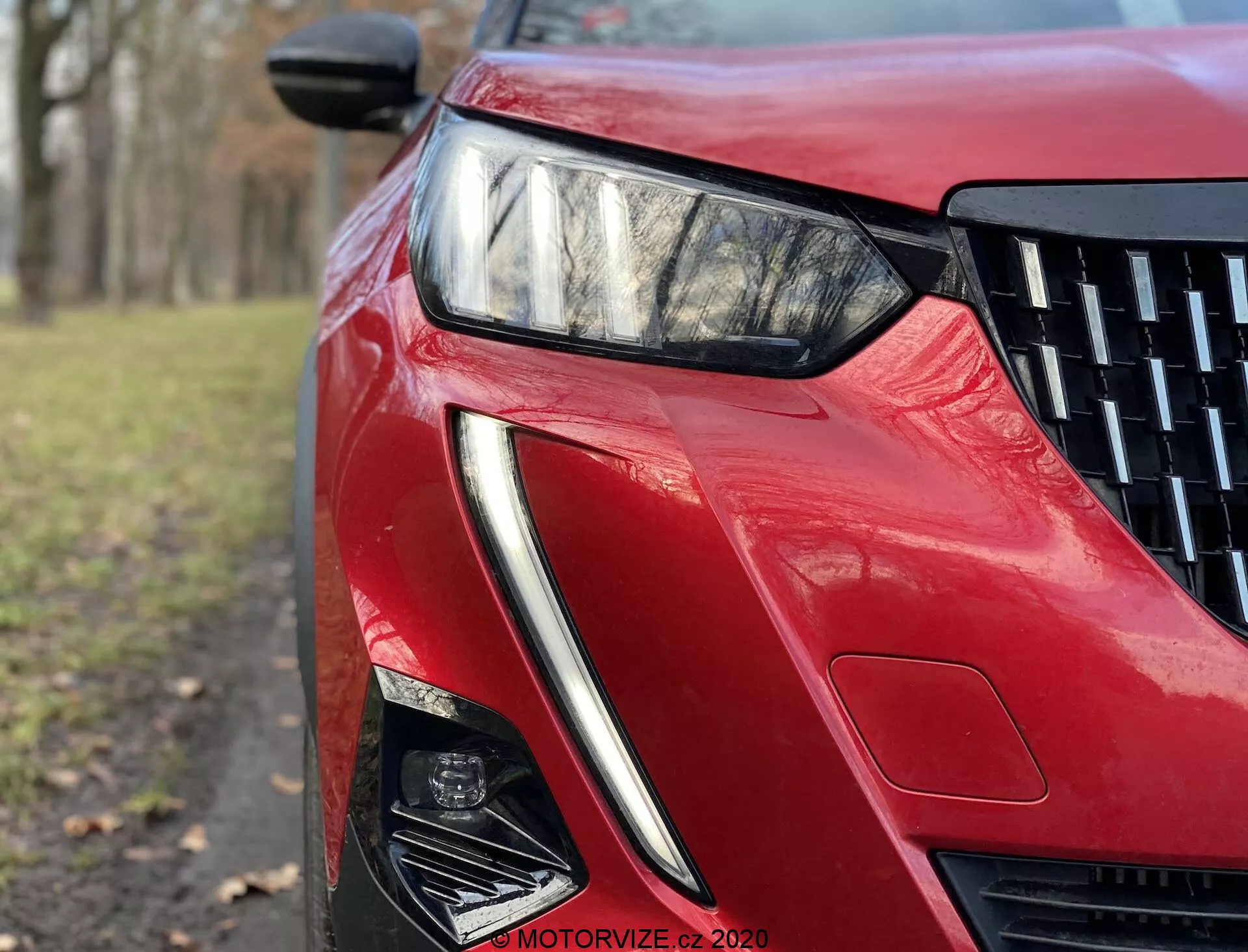 Imagem em close-up de ângulo baixo da seção frontal esquerda do Peugeot 2008 2020 com acabamento vermelho brilhante. A imagem destaca o farol angular de LED, o design nítido da entrada de ar e as linhas esculpidas, refletindo tecnologia avançada de iluminação e estética aerodinâmica.