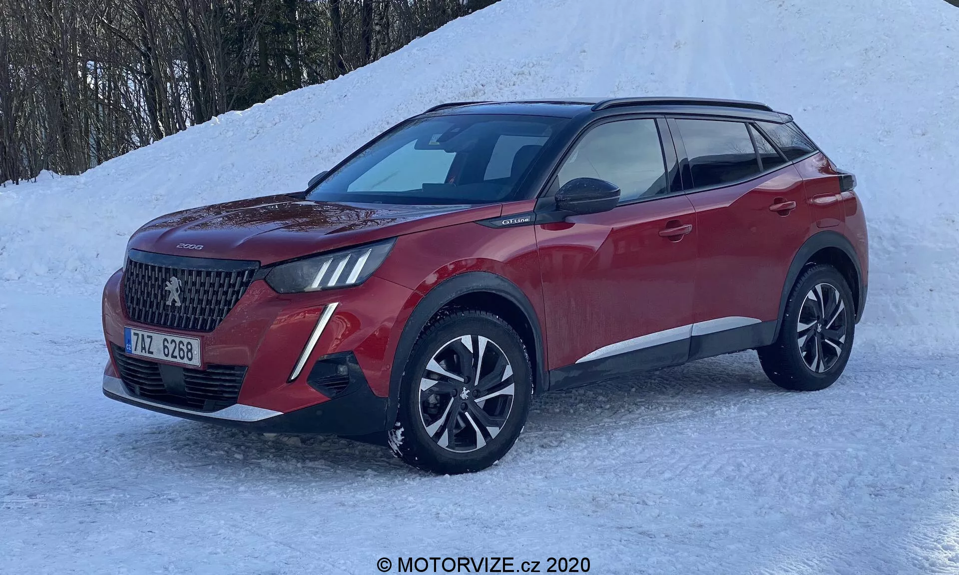 Vista profissional e detalhada de três quartos da frente de um crossover SUV Peugeot 2008 2020 vermelho, capturada do ângulo do lado do passageiro, destacando seu design exterior moderno, grande grade frontal com padrão detalhado, faróis LED estreitos e angulares, faróis de neblina inferiores e uma possível placa de proteção dianteira inferior. A imagem mostra o veículo estacionado em uma superfície nevada, sugerindo suas capacidades de condução no inverno ou em condições de clima frio. Características notáveis incluem rodas de liga leve de múltiplos raios com pneus de inverno ou para todas as estações, espelhos laterais com sinais de direção integrados, linha de caráter dinâmica, trilhos no teto para carga adicional, vidros escurecidos, entrada sem chave e maior altura do solo típica dos SUVs. O veículo está em bom estado e bem conservado, em um ambiente que sugere aptidão para condições de neve ou off-road.