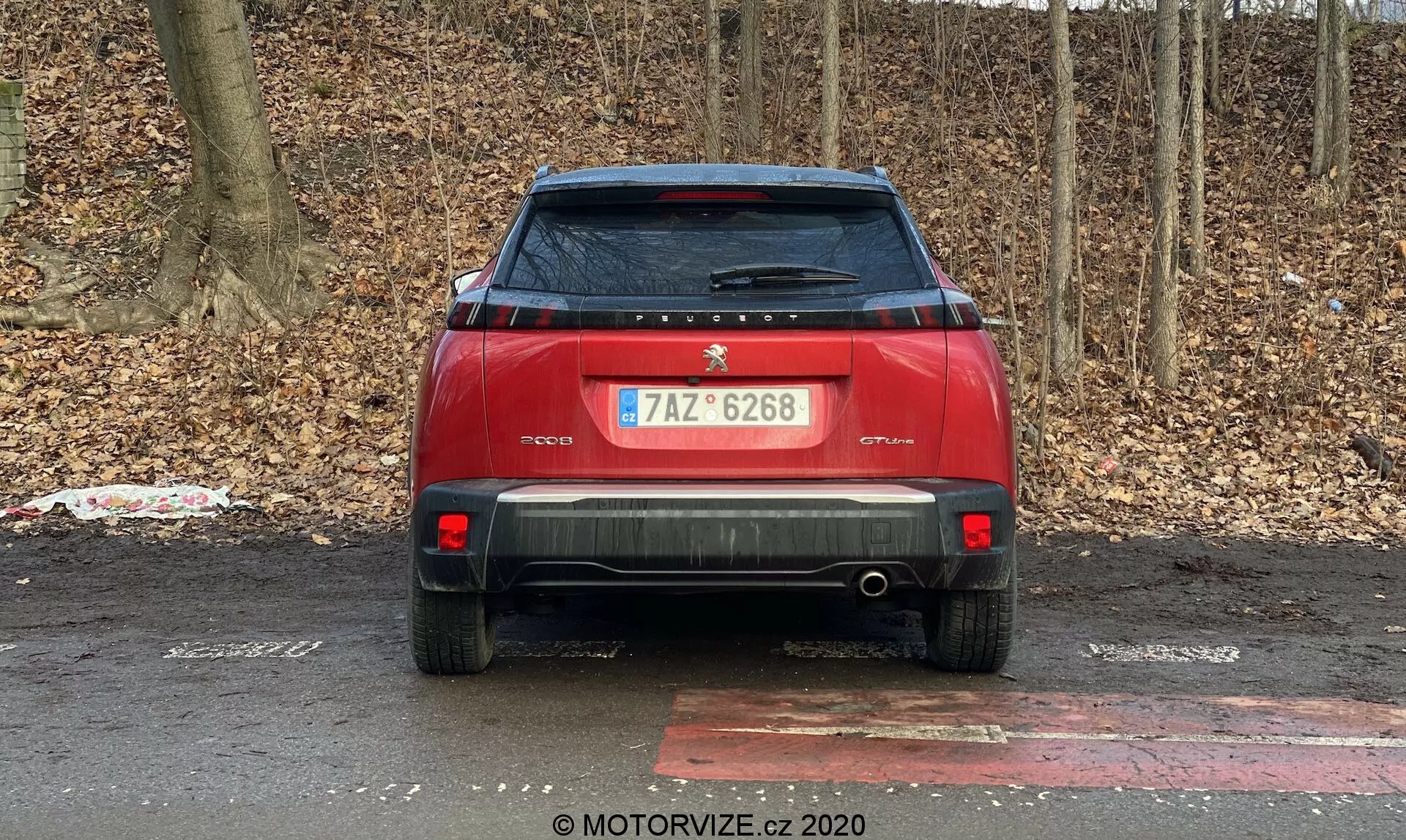 Vista traseira de um carro vermelho estacionado ao ar livre contra um fundo de arbustos ou árvores sem folhas, sugerindo outono ou inverno. As características do carro incluem um logotipo central e placa de identificação na tampa do porta-malas, placa de registro, limpador de para-brisa traseiro, spoiler traseiro, luzes traseiras modernas, refletores e sensores de estacionamento traseiros na área do para-choque, um para-choque traseiro proeminente com um tubo de escape visível, placa de proteção traseira, uma antena de barbatana de tubarão no teto e um adesivo descascando no lado inferior esquerdo da tampa do porta-malas.