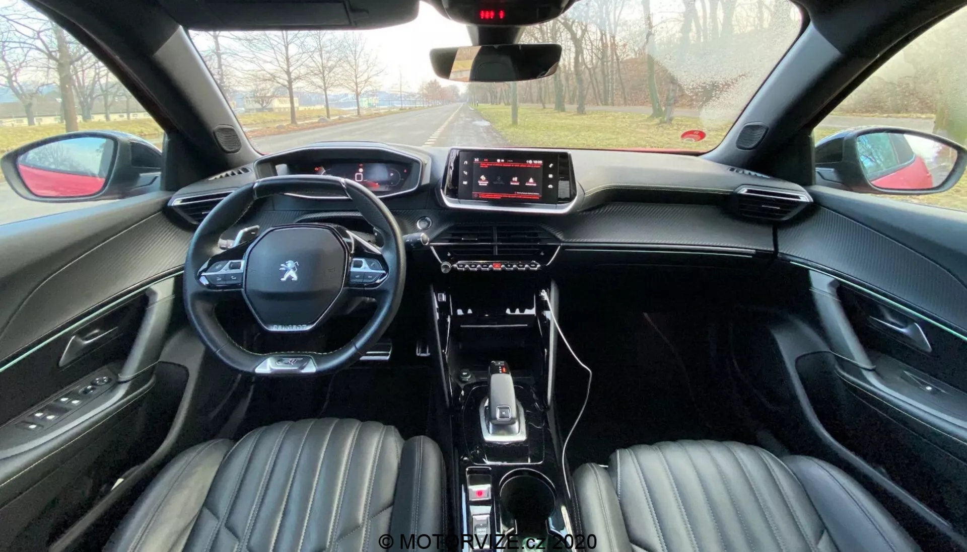 Imagem do interior do Peugeot 2008 2020 (peugeot-2008-1-2-puretech-100-active) da perspectiva do banco traseiro. A imagem mostra os bancos da frente estofados em couro preto com design canelado, apoios de cabeça integrados, um volante multifuncional com o logótipo Peugeot e uma moderna consola central. O painel de instrumentos apresenta um ecrã tátil de infoentretenimento com controlos físicos mínimos, indicando um foco na interação por toque. O carro inclui uma alavanca de câmbio automática, seletores de modos de condução, travão de estacionamento elétrico e compartimentos de armazenamento. A atmosfera sugere um dia claro com uma visão desobstruída da estrada à frente.