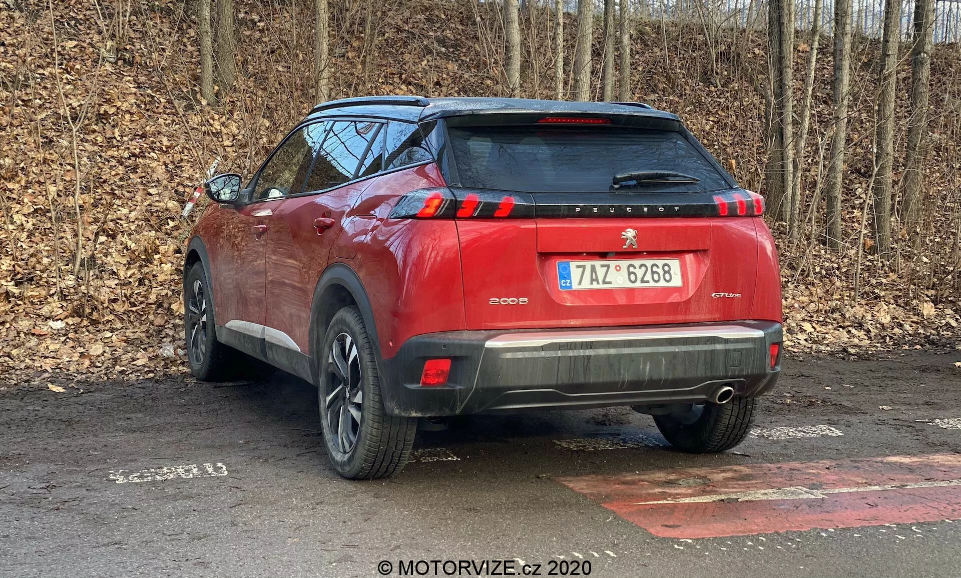 Esta fotografia retrata a vista traseira de um SUV Peugeot 2008 2020 (Peugeot-2008-1-2-PureTech-100-Active) vermelho estacionado ao ar livre. Tirada de um ângulo de três quartos traseiros, mostra o perfil traseiro e lateral do carro, com ênfase em características como as luzes traseiras, para-choques traseiro com potenciais sensores de estacionamento, spoiler traseiro e design parcial das rodas. As modernas luzes traseiras de LED, o esquema de pintura de dois tons, a placa de licença centralizada e os elementos refletivos na parte inferior do para-choques aumentam a segurança e o apelo estético. Situado em uma superfície molhada, coberta de folhas, com árvores ao fundo, indicando um ambiente não urbano, a fotografia captura detalhes finos como a textura do material, a clareza do vidro e o design da banda de rodagem do pneu.