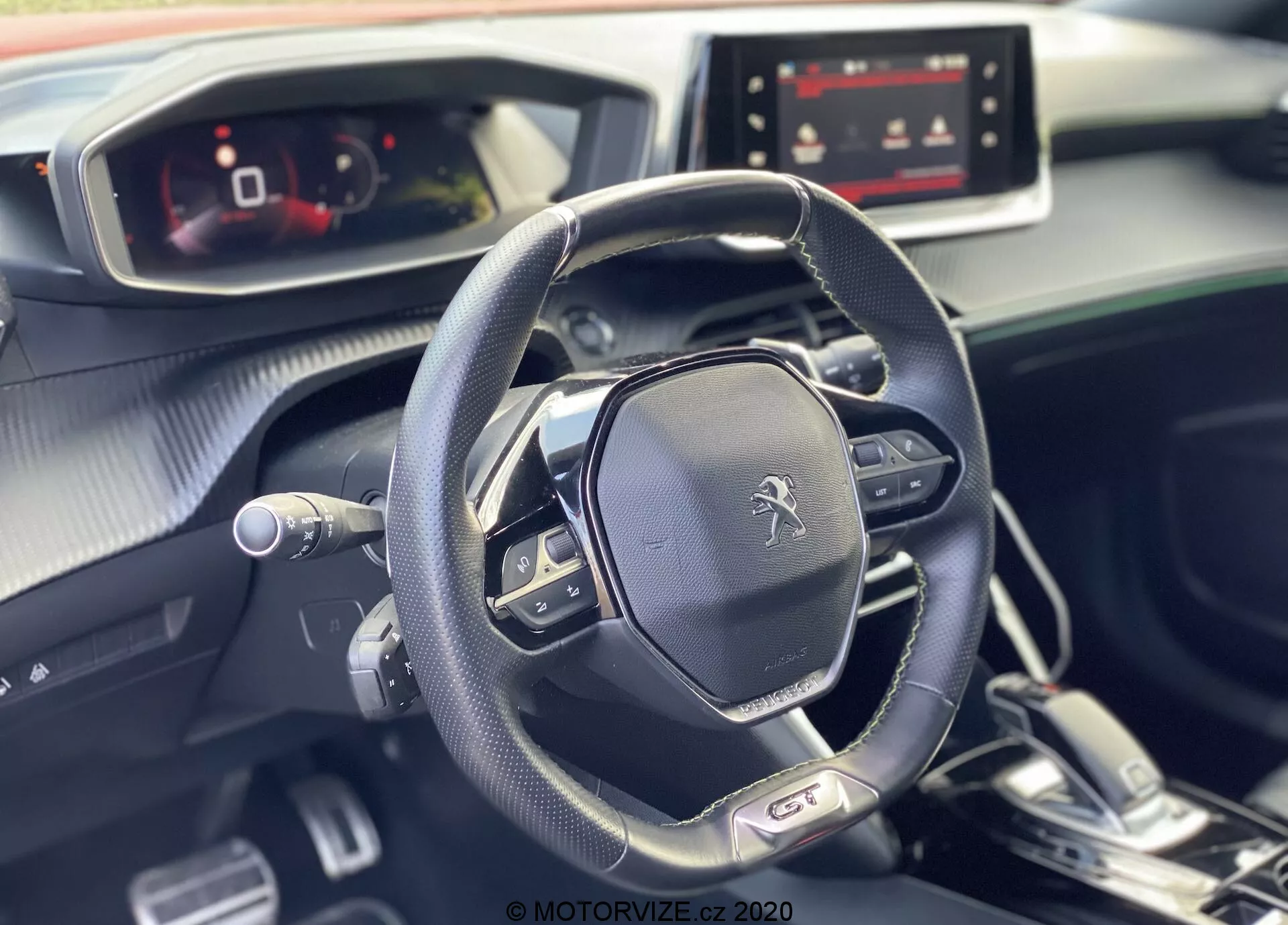 A imagem mostra o interior moderno e elegante do Peugeot 2008 2020, destacando o volante multifuncional e o painel de instrumentos totalmente digital a partir da perspectiva do passageiro. O emblema Peugeot é exibido de forma proeminente no volante, que possui controles de mídia, configurações de controle de cruzeiro e aletas de mudança, sugerindo uma transmissão automática com opções de mudança manual. O painel, projetado com uma combinação de materiais suaves ao toque e acentos metálicos, inclui uma tela de infoentretenimento ligada, sugerindo várias opções de conectividade e navegação. A luz natural realça a atmosfera impecável e agradável do interior do carro.