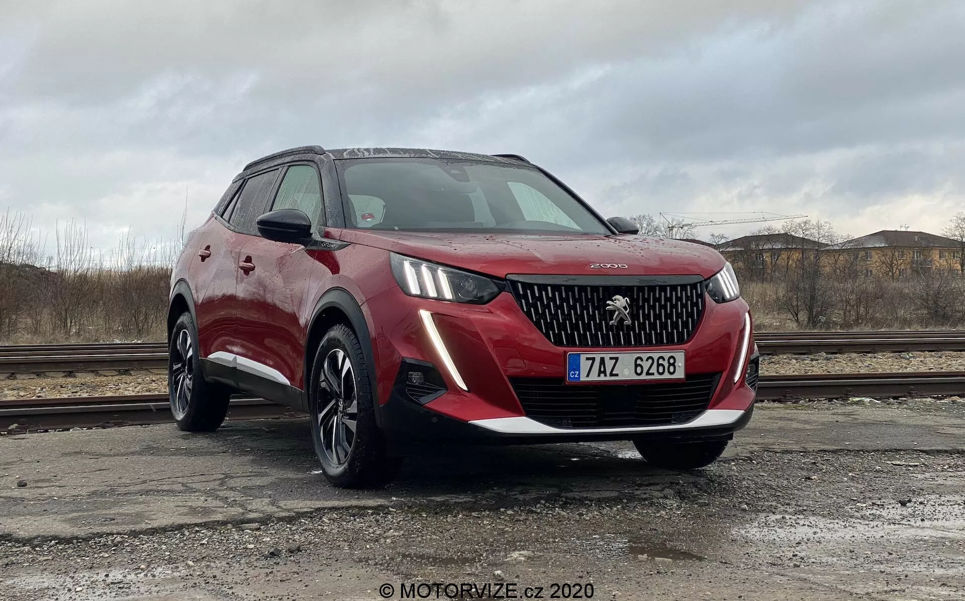 Um SUV Peugeot 2008 2020 (Peugeot-2008-1-2-PureTech-100-Active) vermelho estacionado ao ar livre contra um céu nublado com terreno árido e entulho de construção ao fundo. O veículo é capturado de um ângulo baixo e próximo de três quartos frontais, mostrando a grade frontal com o logotipo da Peugeot, elegantes faróis de LED, luzes de nevoeiro, rodas de liga leve multirraiadas e um acabamento de pintura metálica vermelha com revestimento de plástico preto em torno dos arcos das rodas. Os espelhos laterais, parcialmente visíveis, parecem ser da cor da carroçaria com indicadores de direção integrados, e o pára-brisas inclinado proporciona boa visibilidade para o condutor. As características prováveis incluem sensores de estacionamento e câmaras, inferidas do seu design contemporâneo.