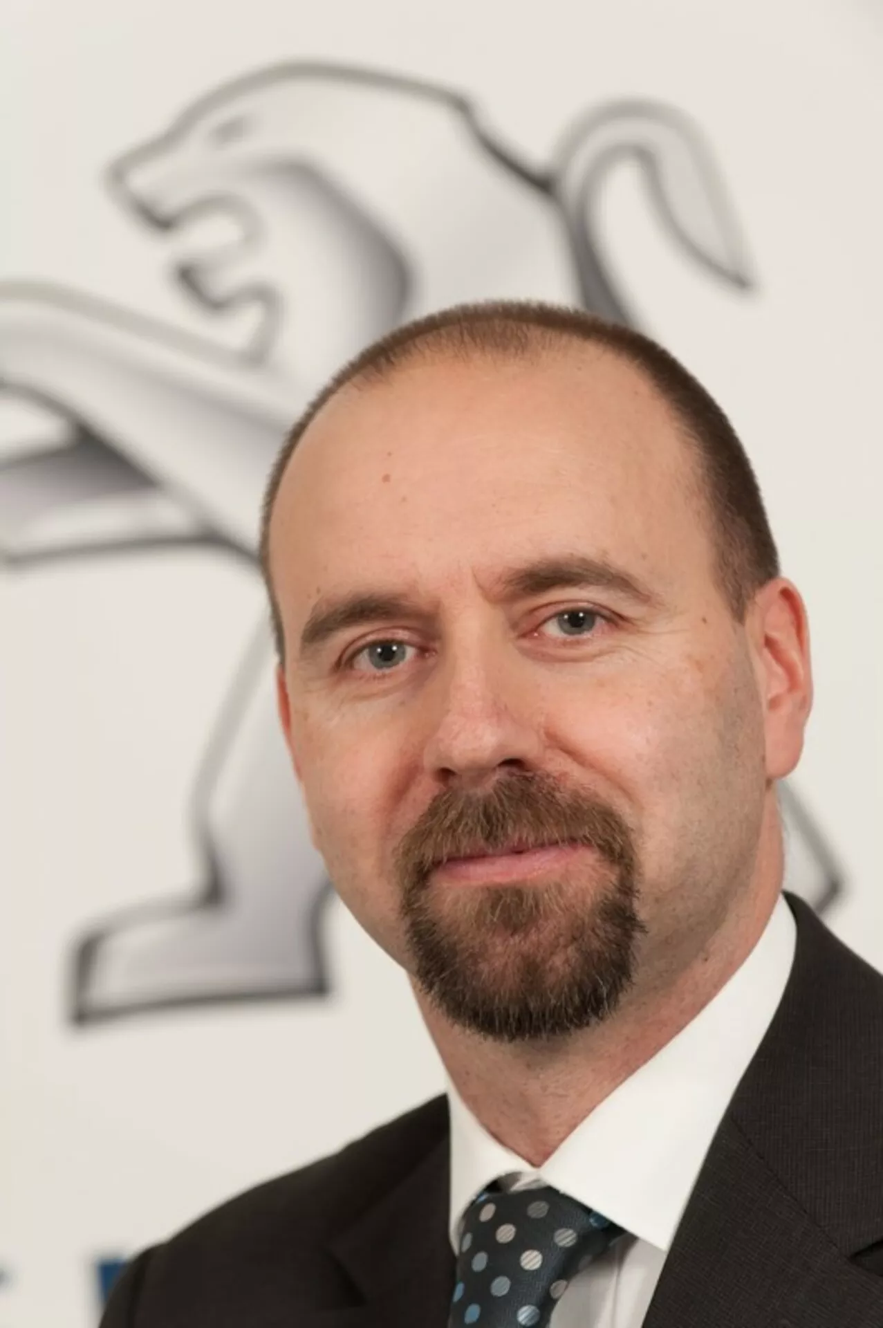 Retrato profissional de um homem de terno e gravata com o logotipo do leão da Peugeot ao fundo, sem mostrar um carro