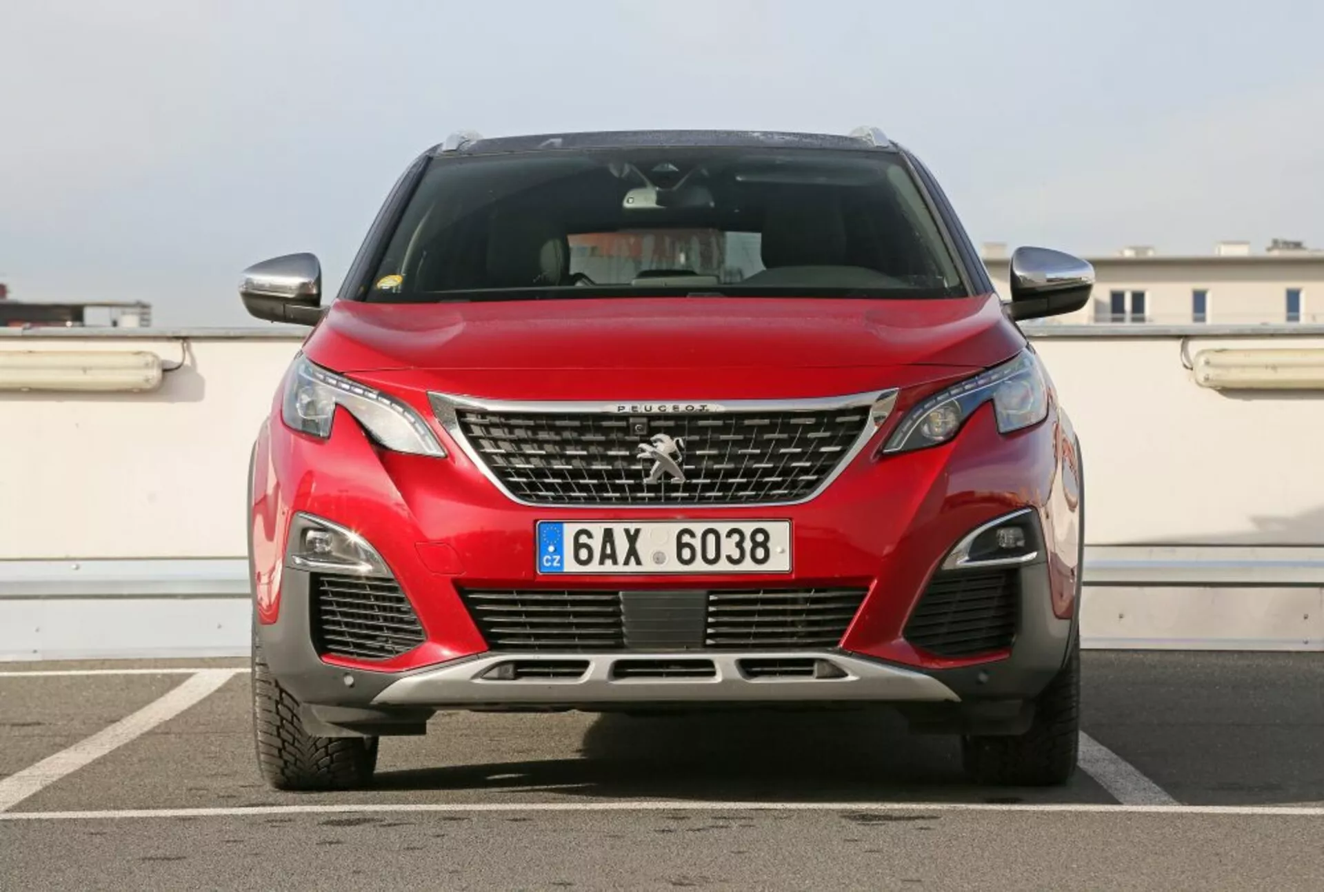 Vista frontal de um Peugeot 3008 vermelho de 2016 mostrando o emblema na grade, faróis angulares, entradas de ar inferiores com luzes de nevoeiro, vislumbre dos contornos do capô contra um pano de fundo urbano com céu limpo