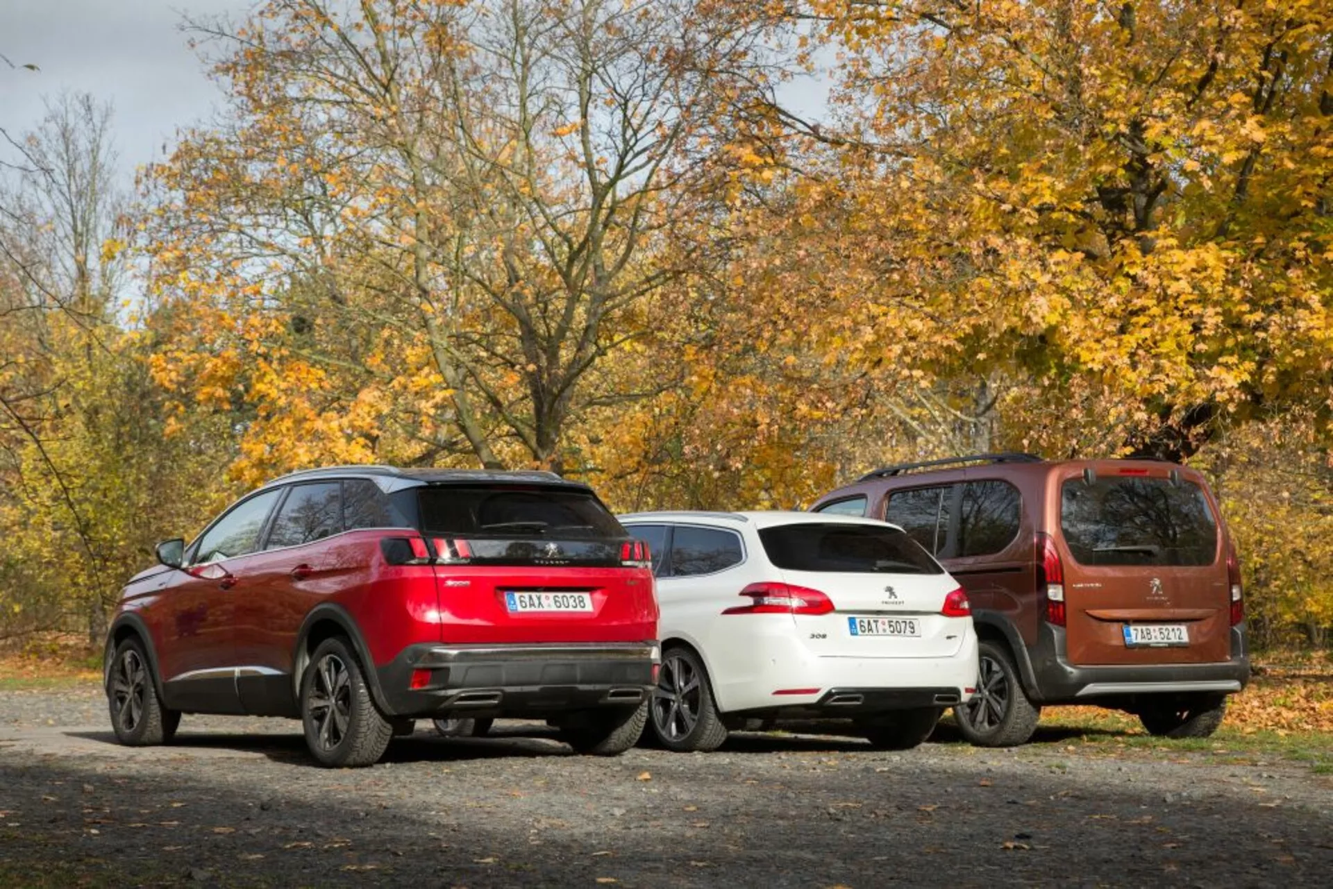 Vista traseira de três carros estacionados em uma superfície de cascalho temática de outono, apresentando um SUV esportivo vermelho, um hatchback branco elegante e um MPV compacto marrom, todos equipados com características modernas como rodas de liga e sensores de estacionamento traseiros, sob luz natural do dia.